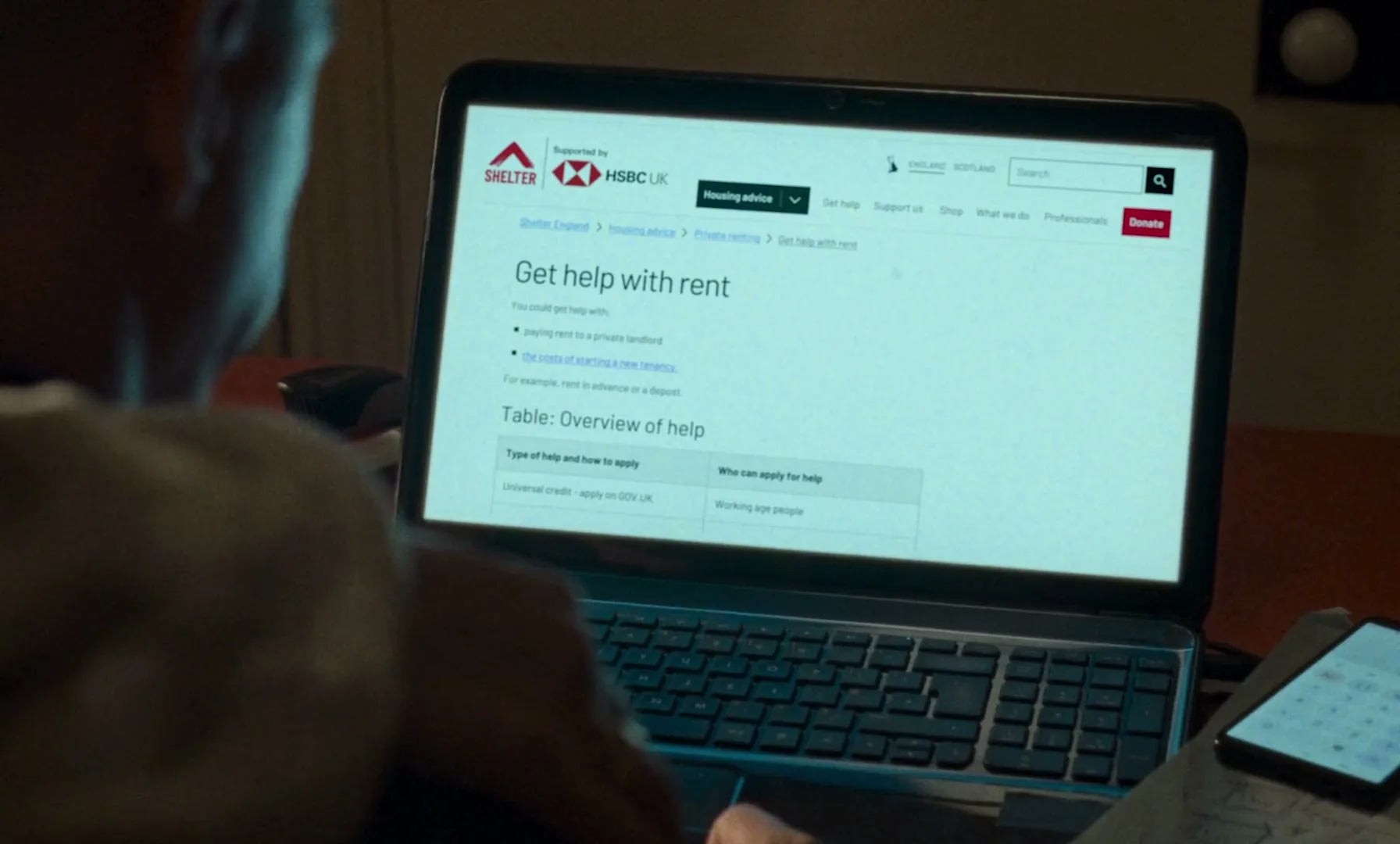 A person sits at a desk, looking at a laptop screen displaying the Shelter website with information about getting help with rent.