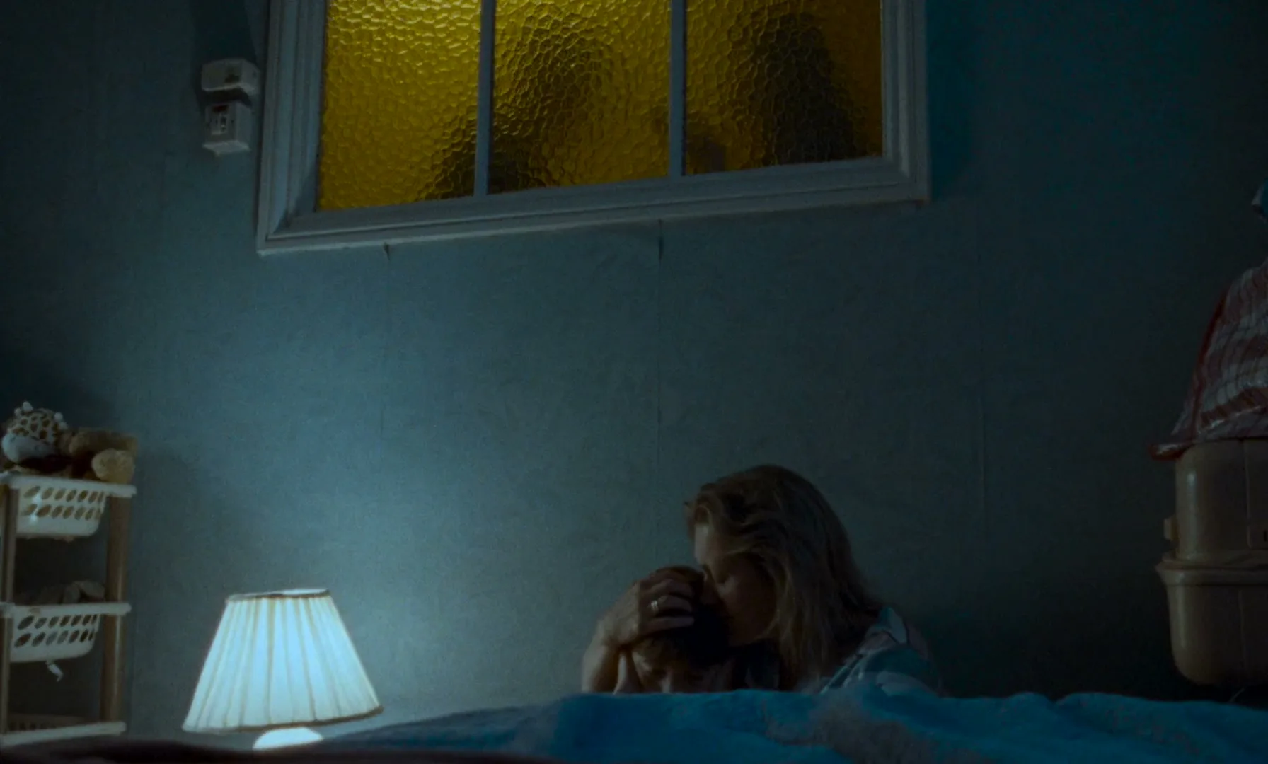 A mother comforts her child in a dimly lit bedroom, with silhouettes visible through a textured window above them.