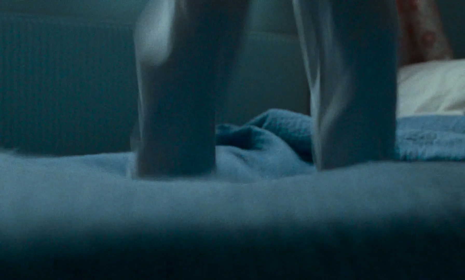 A child's feet in blue socks jump up and down on a bed with blue sheets in a dimly lit bedroom.