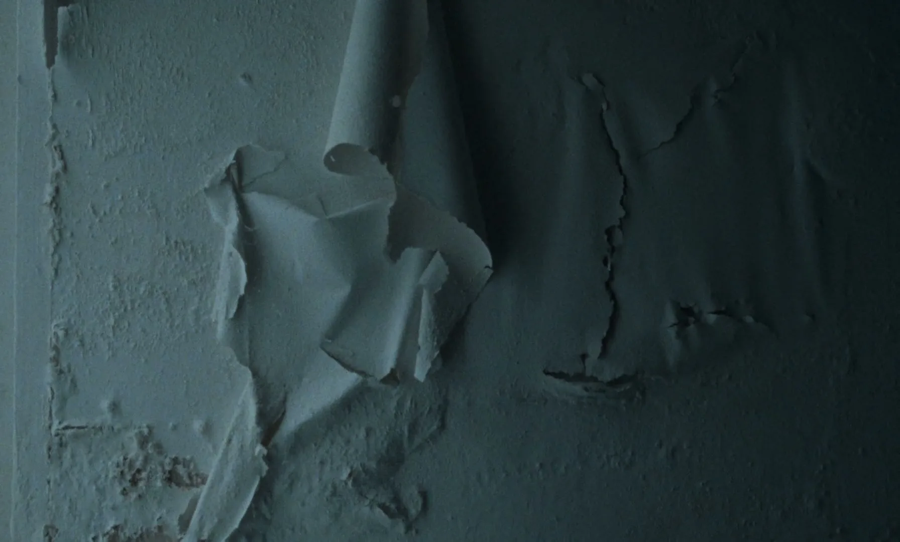 Close-up of peeling paint on a wall, revealing damaged plaster underneath, in a dimly lit room.