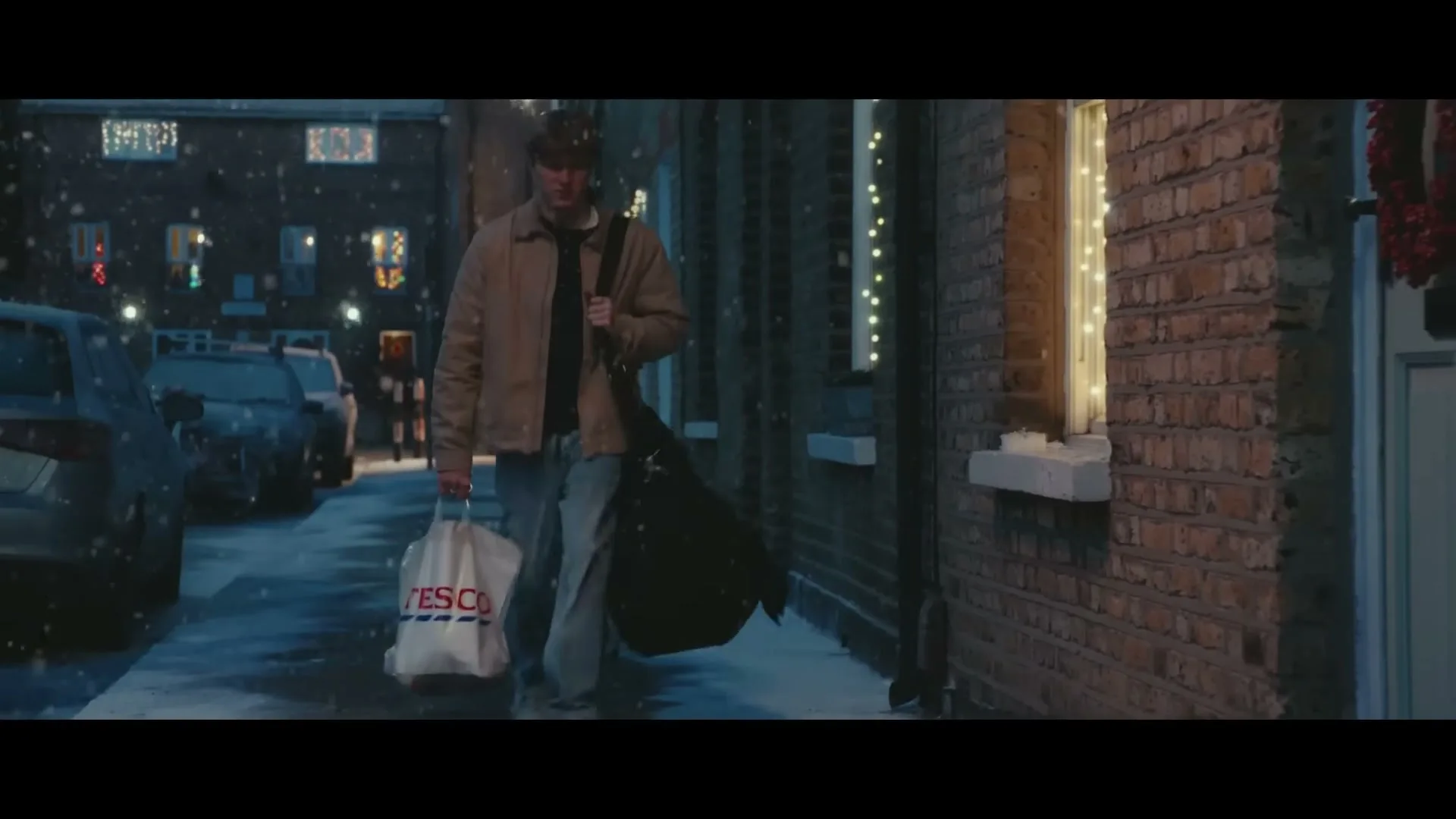 A young man walks down a snowy street at night, carrying a Tesco bag and a duffel bag, with Christmas lights decorating the houses.