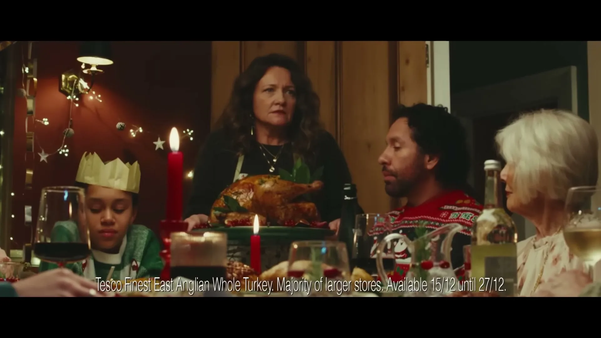 A woman in a black apron places a roasted turkey on a festive Christmas dinner table, surrounded by family members.