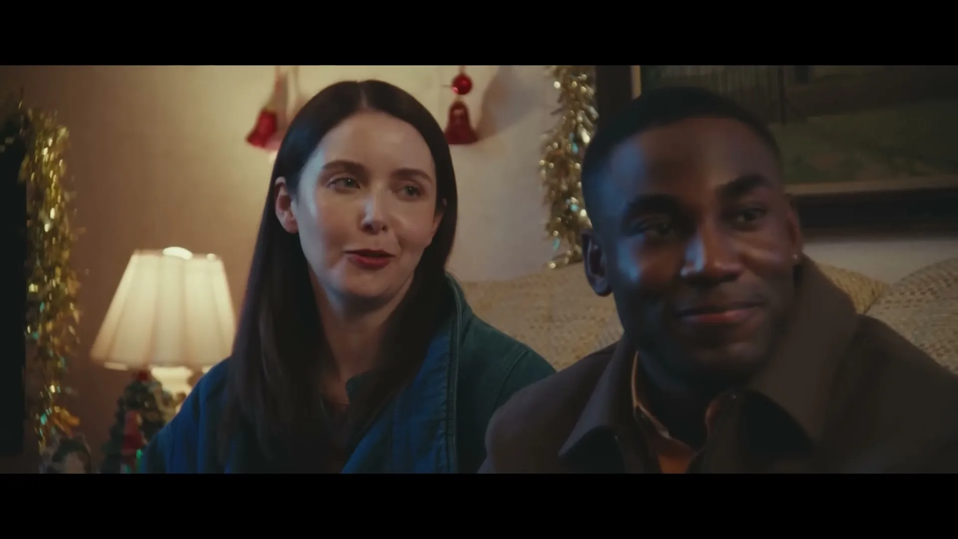 A young white woman and a young Black man sit on a couch in a festive living room, looking off-camera and smiling.