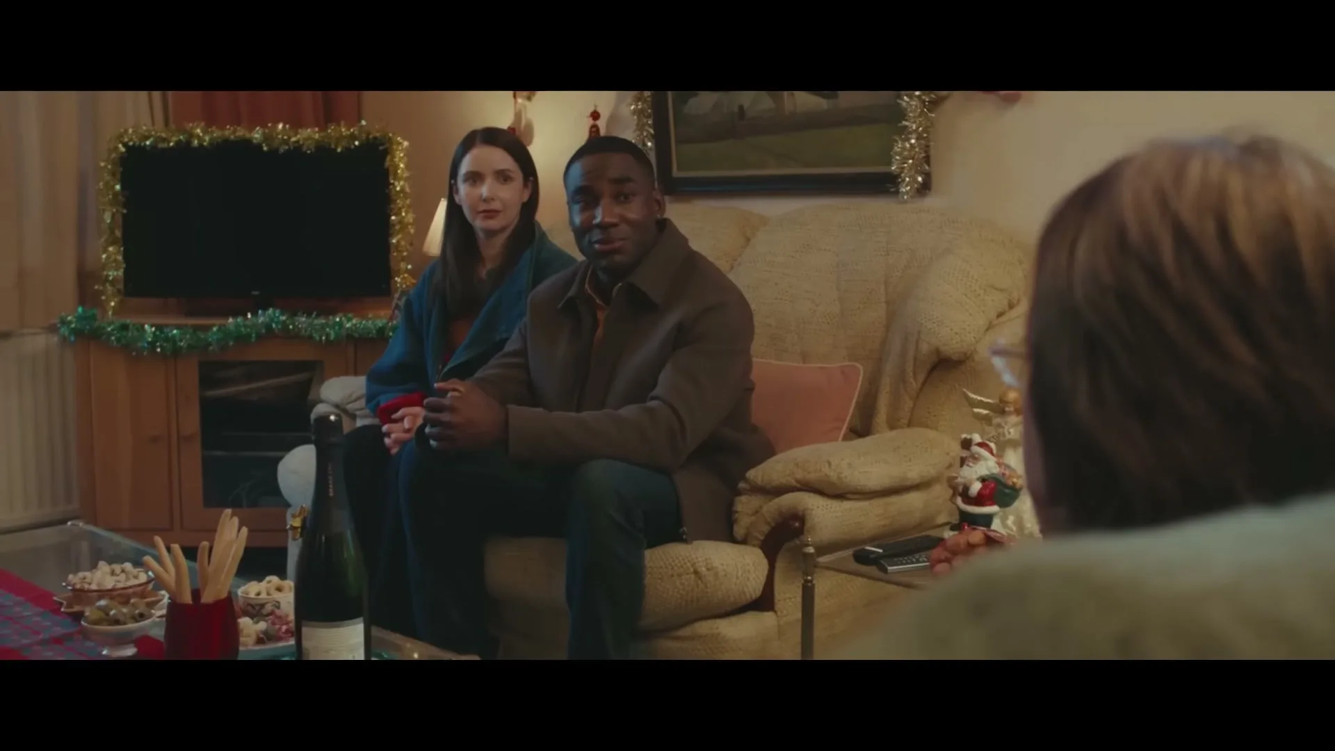 A young couple, a man and a woman, sit on a sofa in a decorated living room, looking towards an unseen person.
