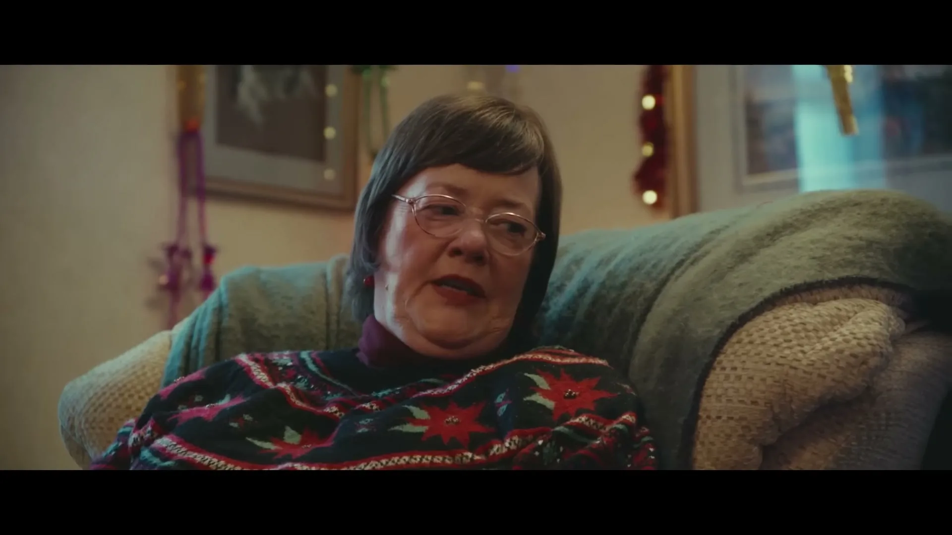 An older woman with glasses and a festive sweater sits on a couch, talking and smiling in a decorated living room.