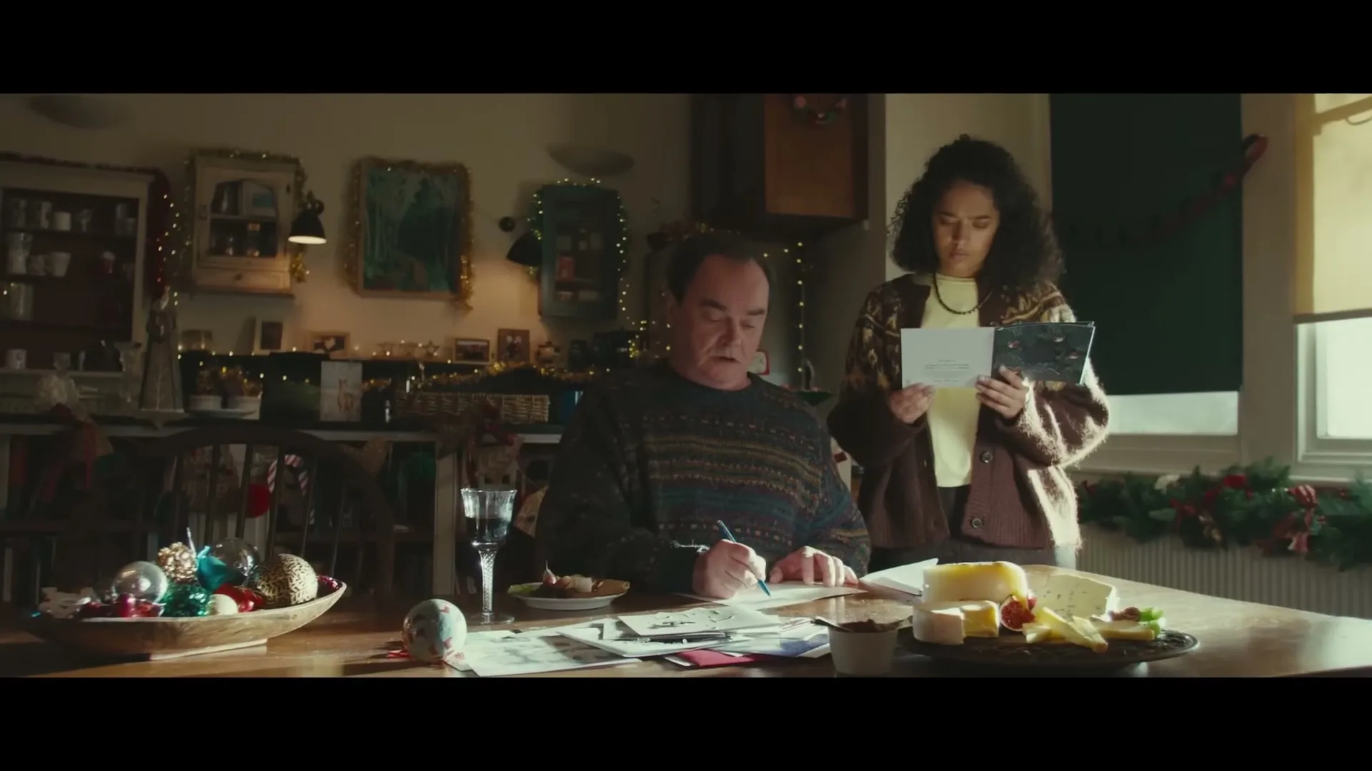 A man sits at a table writing Christmas cards while a woman stands next to him, reading a card, in a festive home.