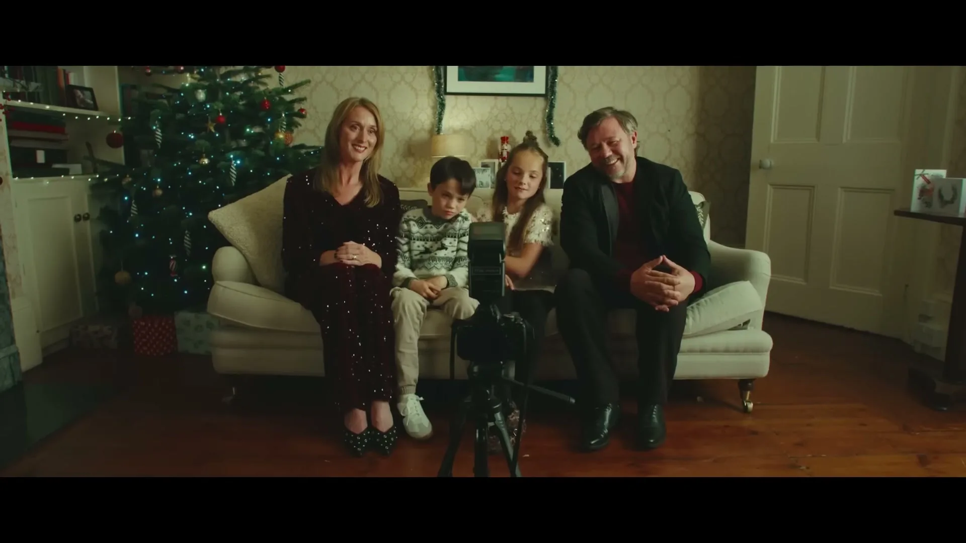 A family of four, a mother, father, and two children, sit on a couch in their living room, posing for a photo in front of a Christmas tree.