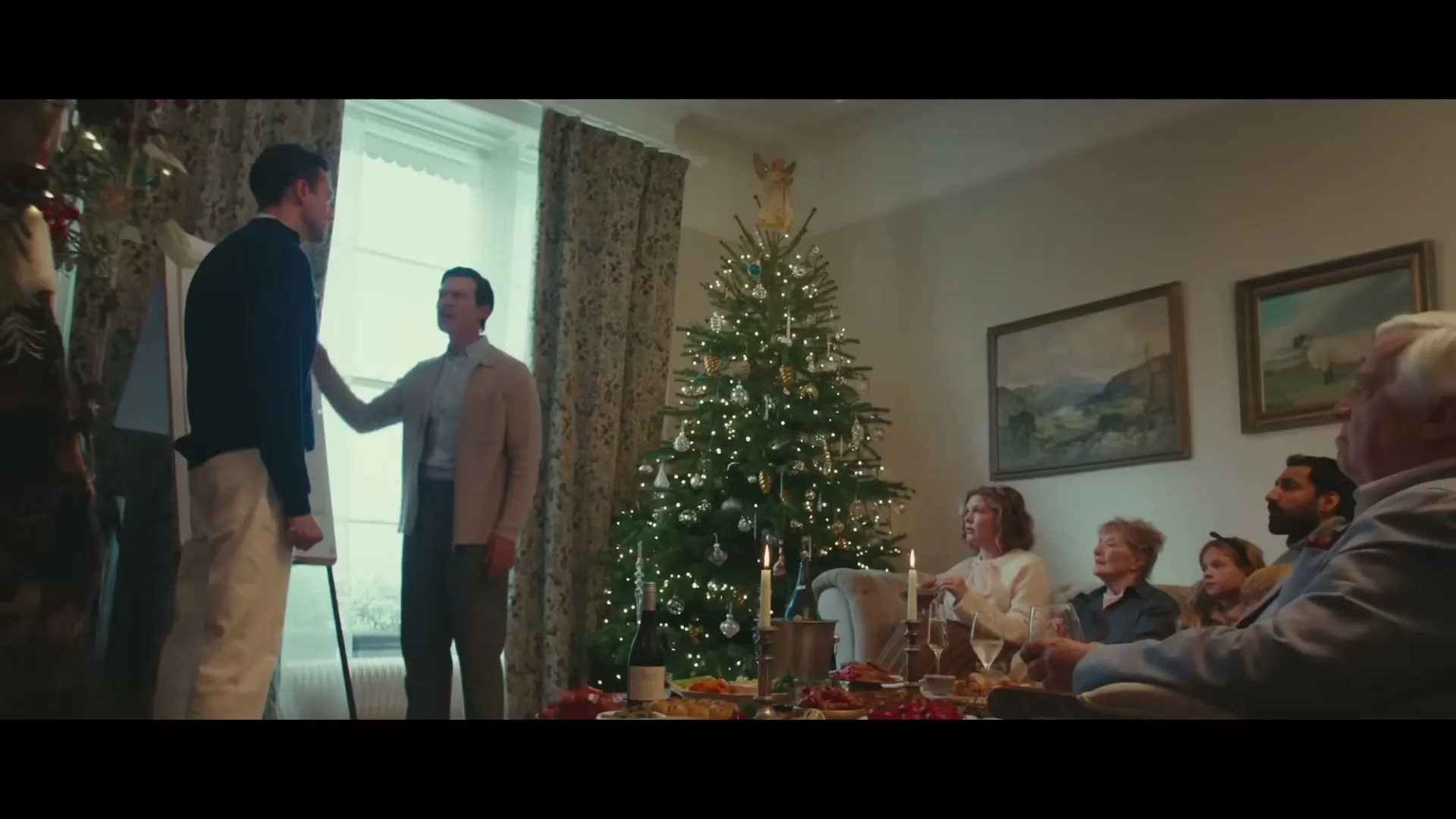 A family gathers around a dining table in a living room, with two men standing and talking near a Christmas tree.