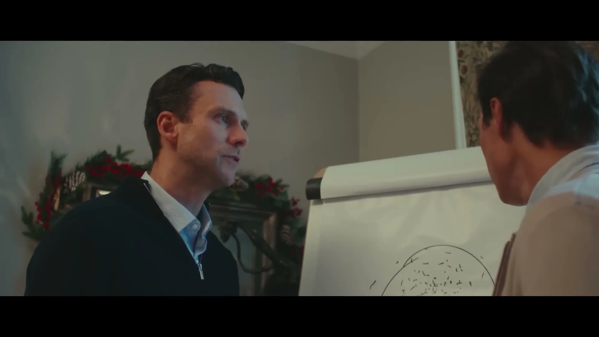 Two men in a living room discuss a drawing on a flip chart, with Christmas decorations visible in the background.