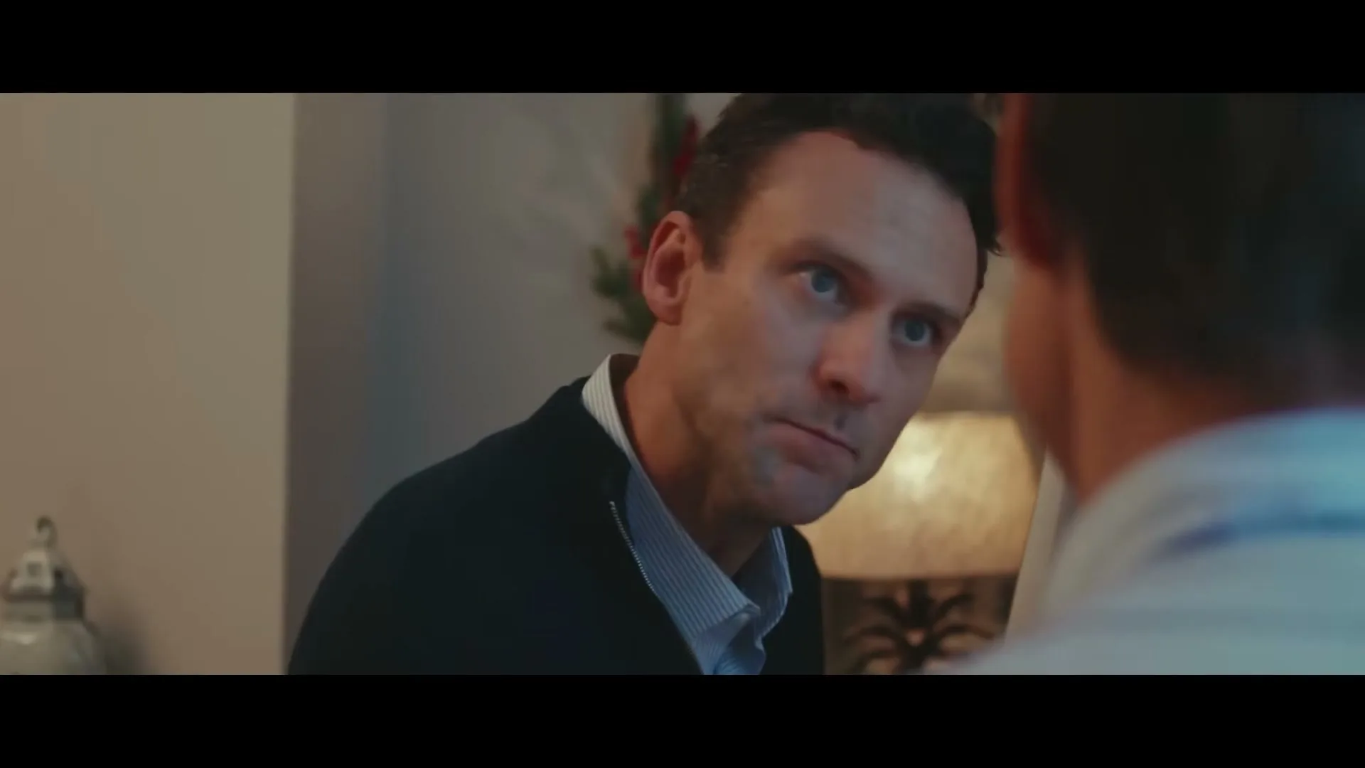 A man in his 30s, wearing a dark blue cardigan over a striped shirt, looks up and to the right, talking to someone off-screen in an interior setting.
