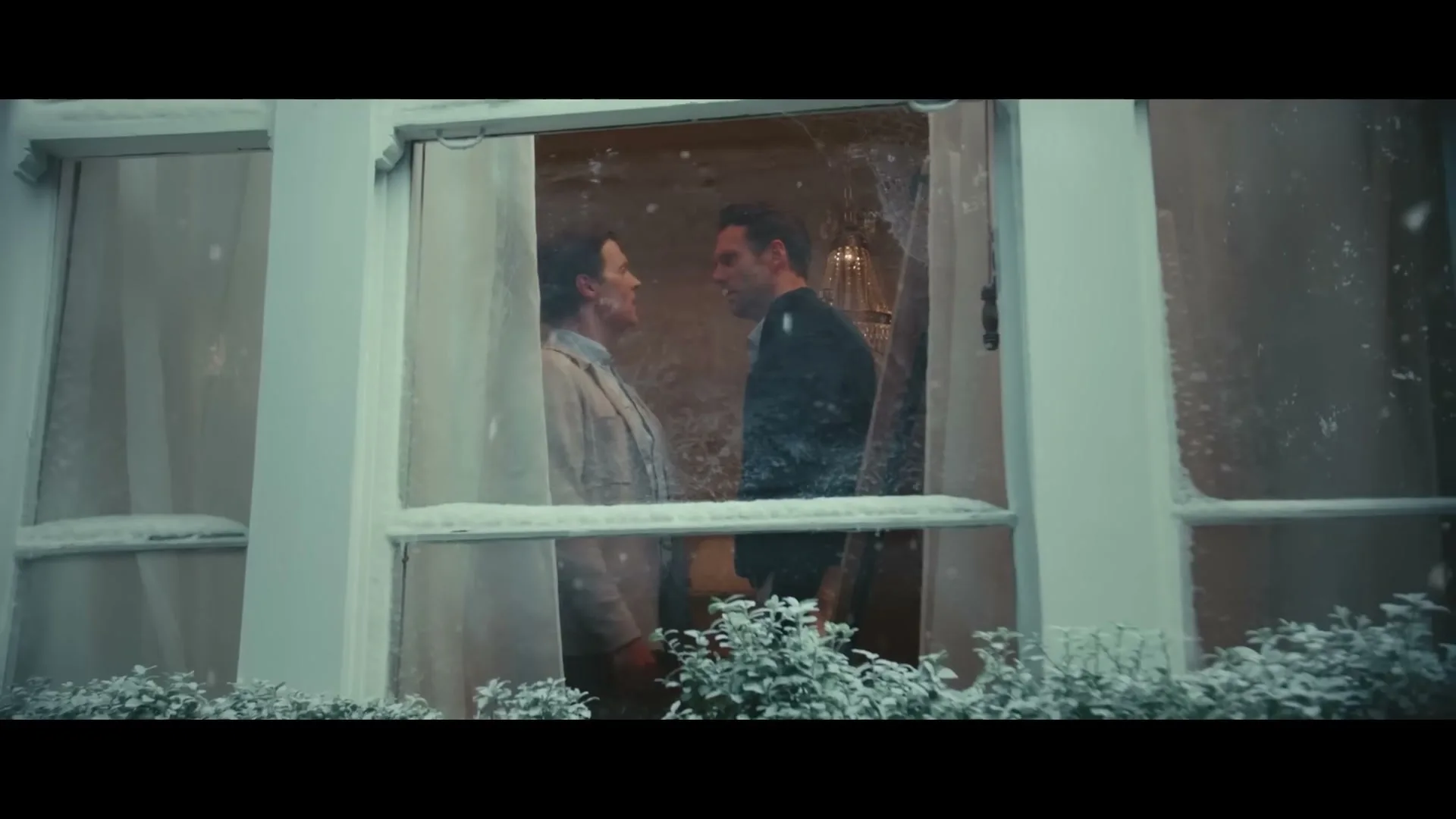 A man and a woman stand close, looking at each other through a snowy window, as snow falls outside a house.