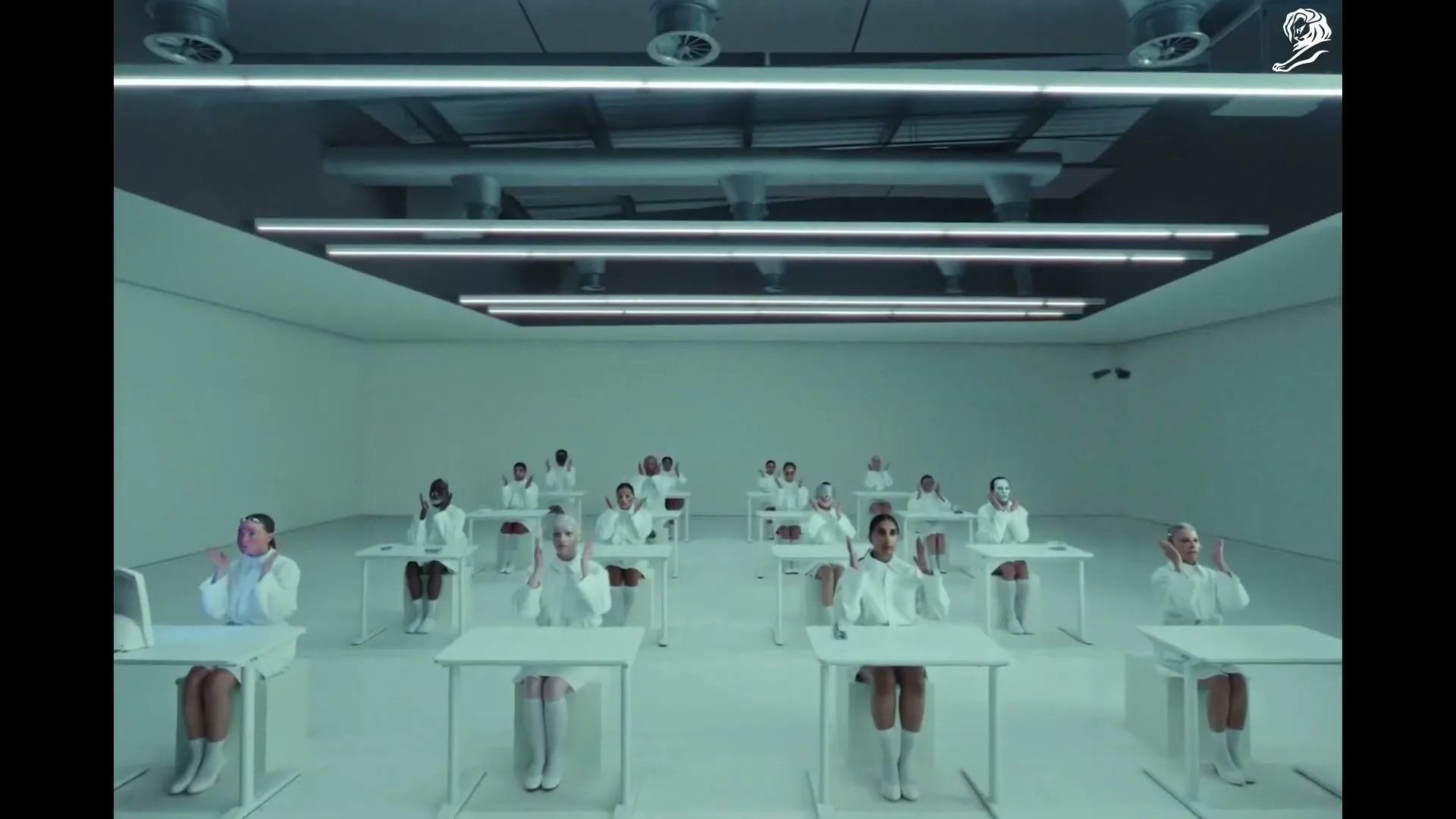 A group of diverse people in white lab coats sit at individual desks in a sterile, white room, performing synchronized hand movements.