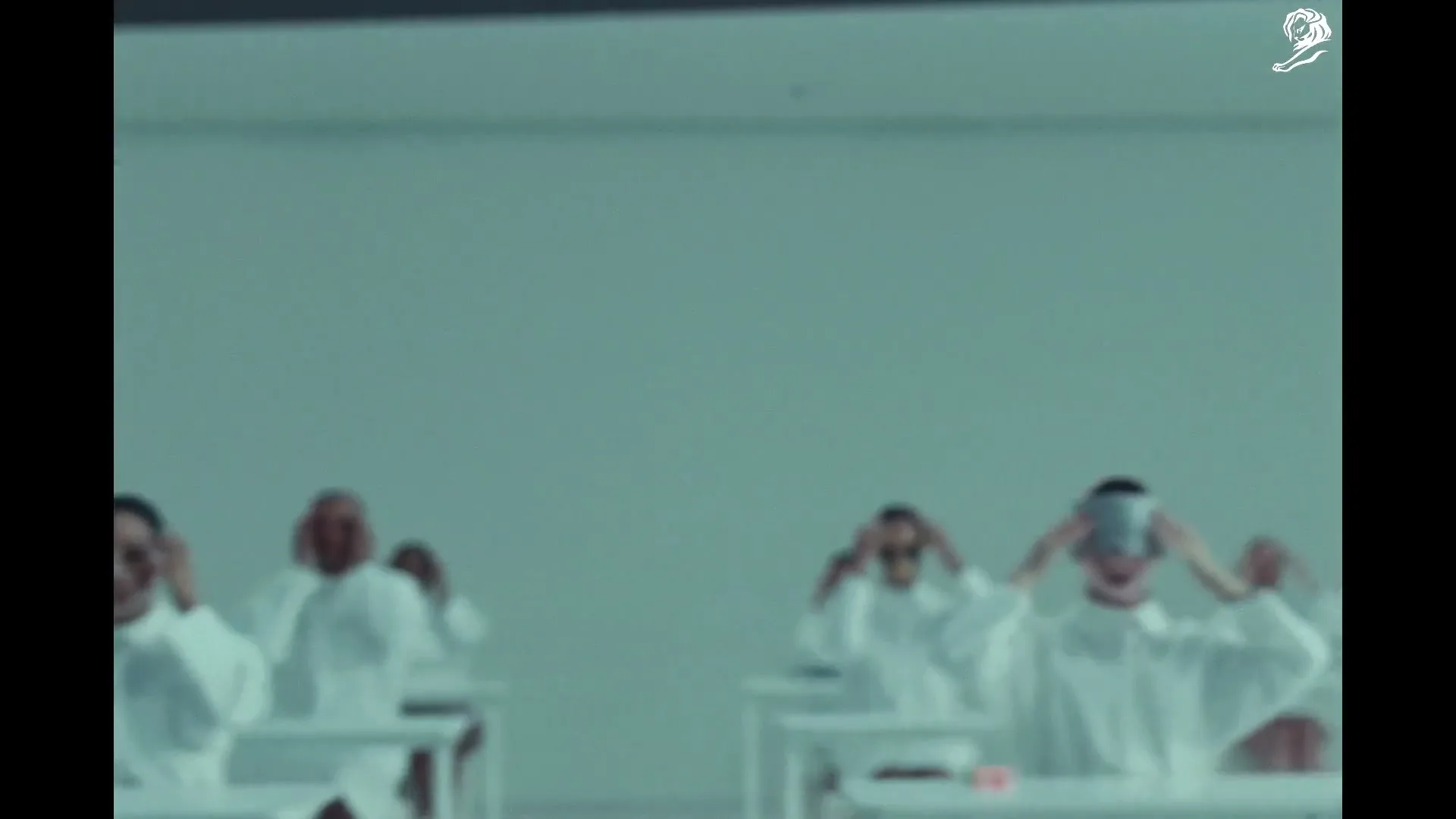 A group of people in white lab coats are seen in a sterile, white room, some with clear masks on their faces, others with silver masks, as they perform synchronized movements.