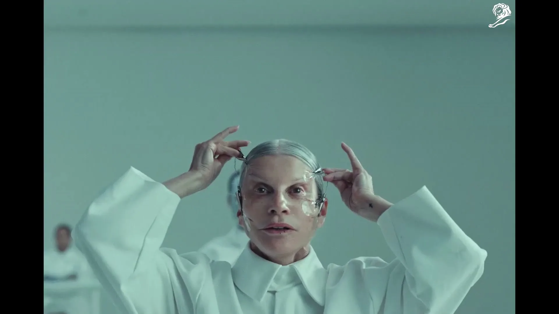 A woman with pale skin and gray hair, wearing a white lab coat, adjusts a futuristic device on her face in a sterile, white room.