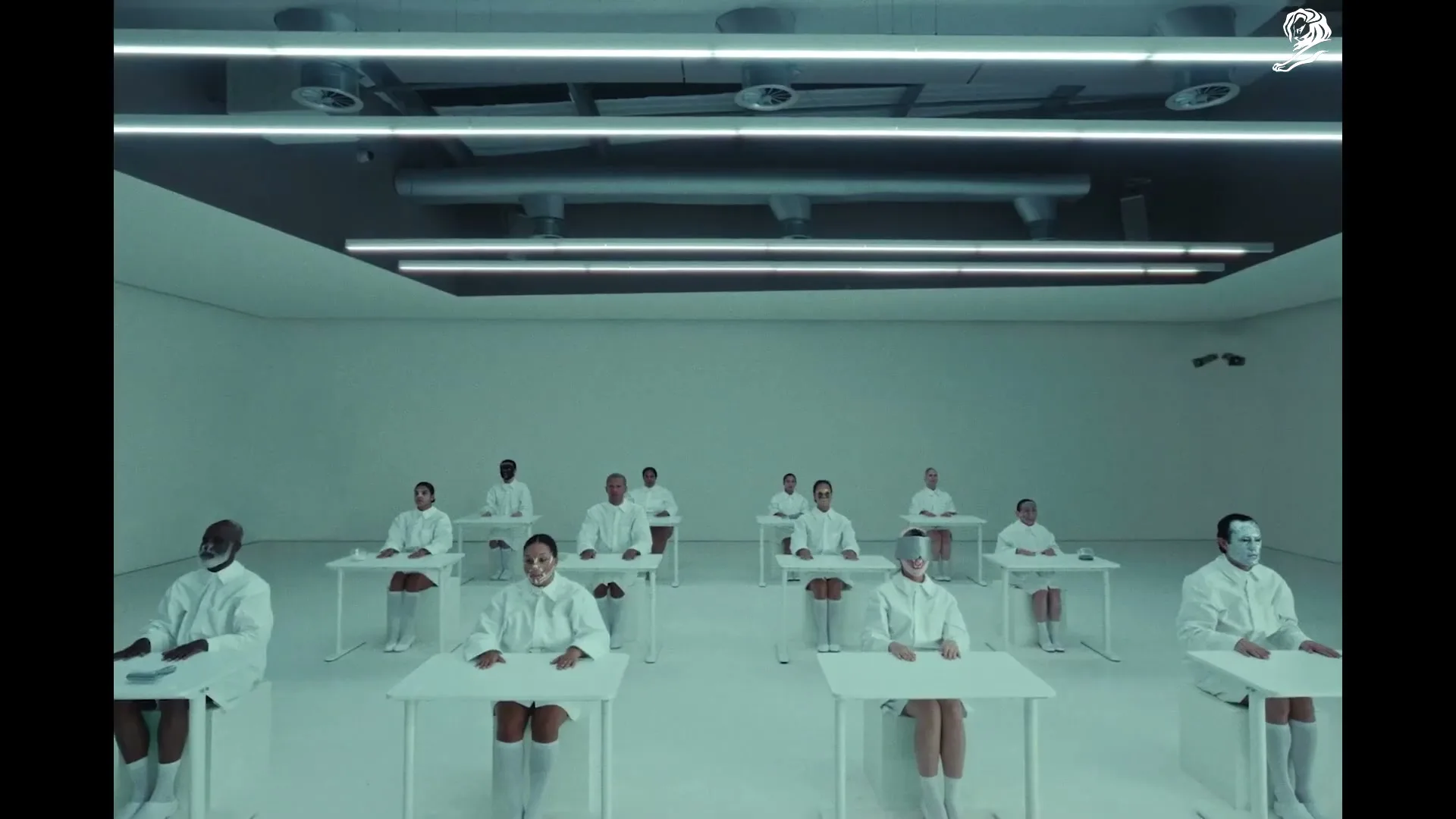 A group of people in white lab coats and masks sit at individual desks in a sterile, white room, looking forward.