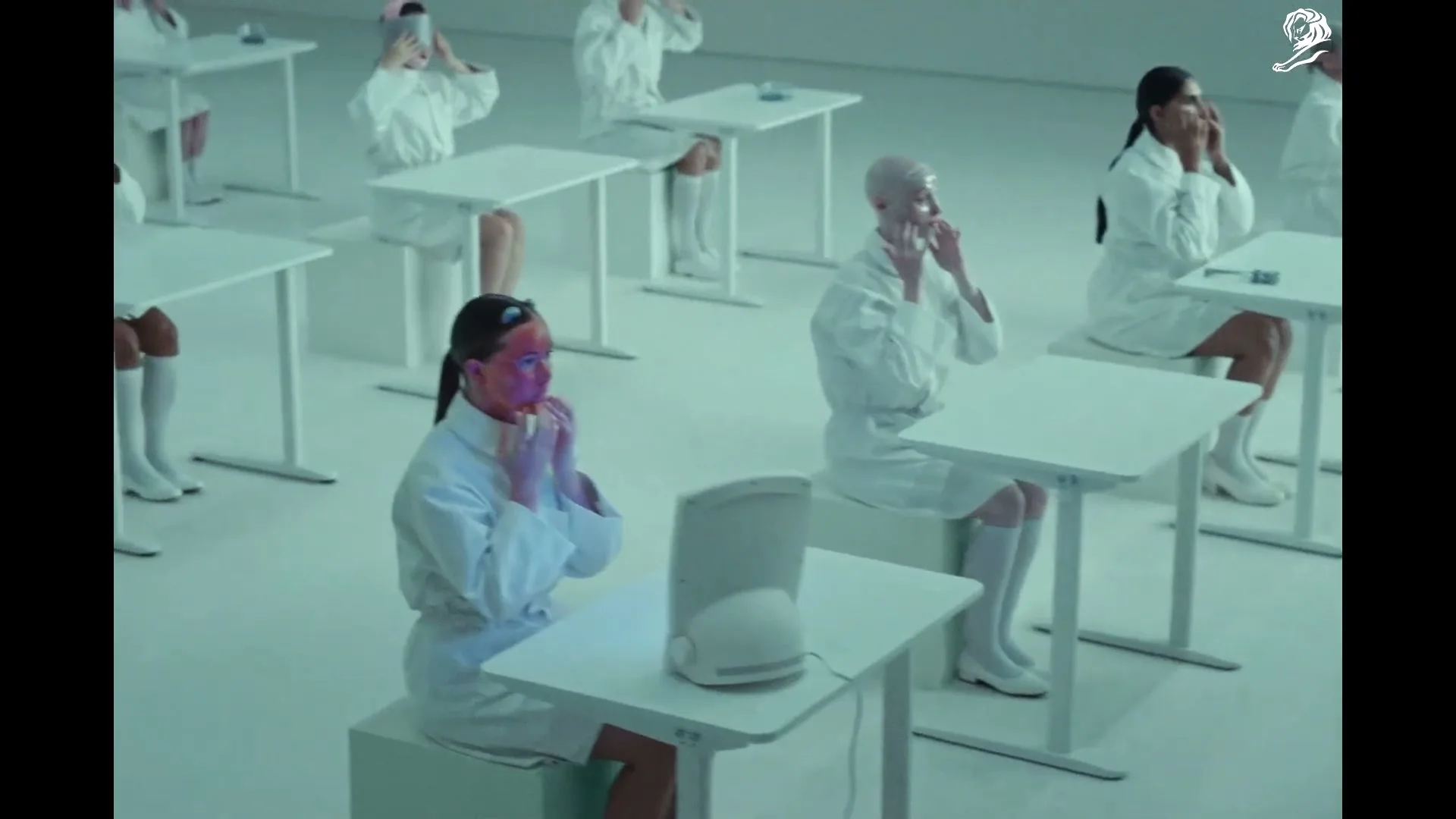 A group of women in white lab coats sit at individual desks, applying face masks in a sterile, futuristic room.