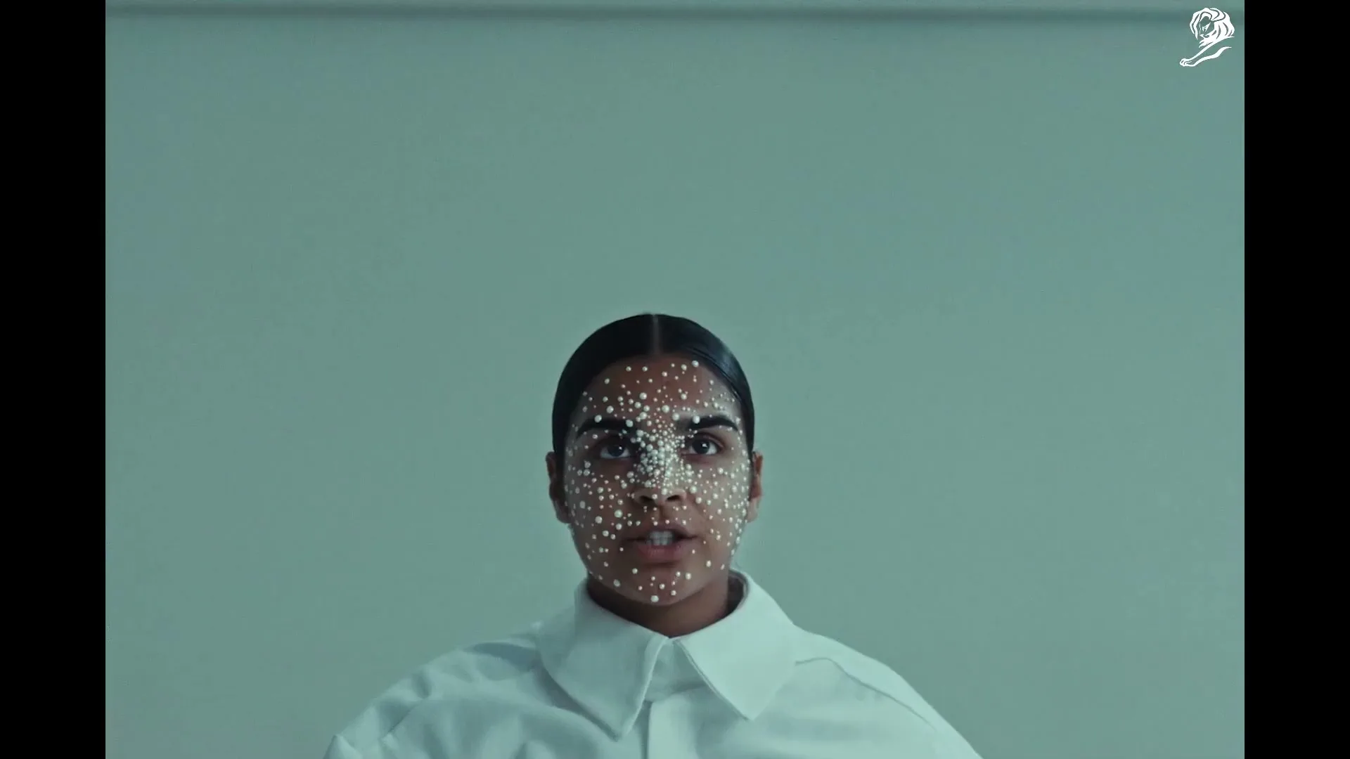 A woman with dark hair and white dots on her face looks directly at the camera, wearing a white collared shirt in a studio.