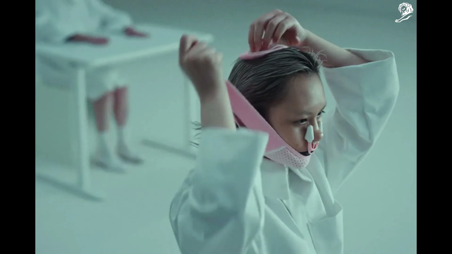 A person with short hair and a white lab coat adjusts a pink facial device with a nose clip in a sterile, white room.