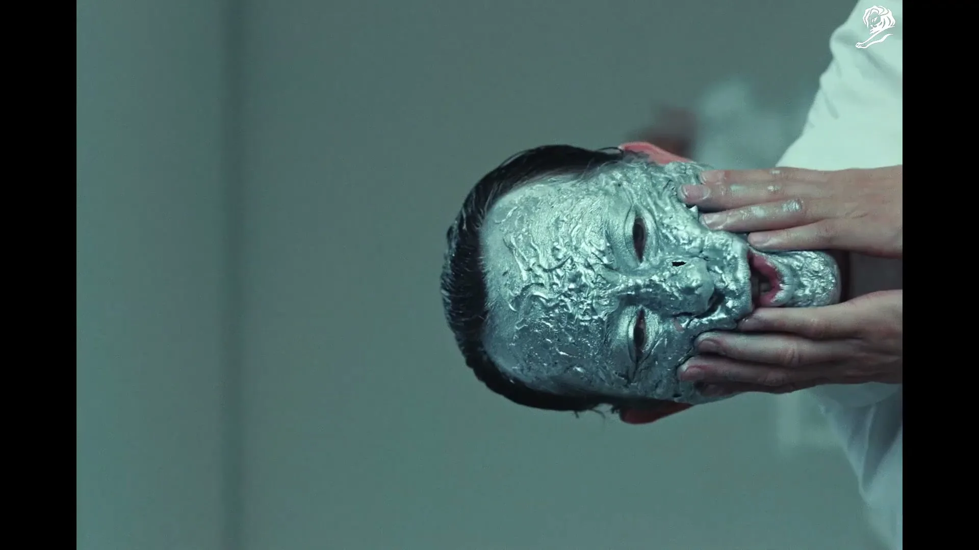 A person with a silver-painted face is being touched by hands in white lab coats, creating a surreal and unsettling image.