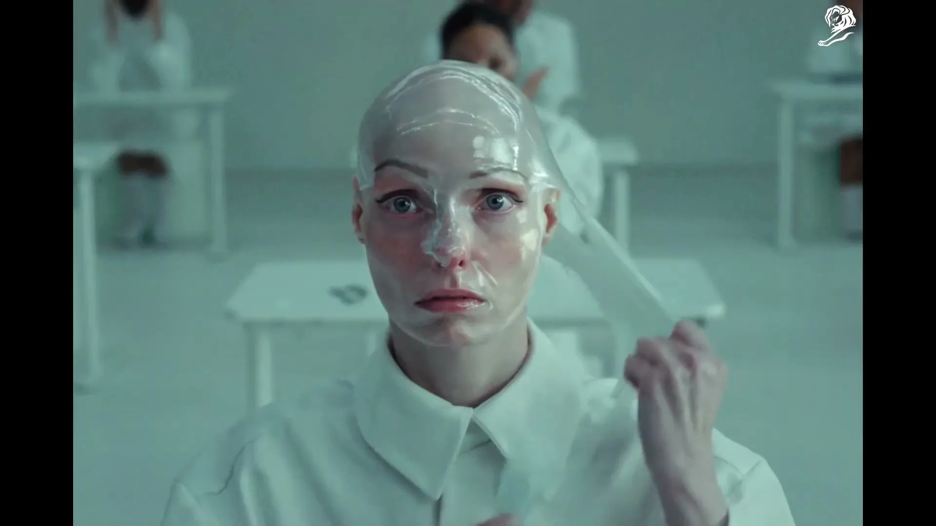 A woman with a bald cap and a clear plastic film on her face looks directly at the camera, pulling the film off her head in a sterile, white room.