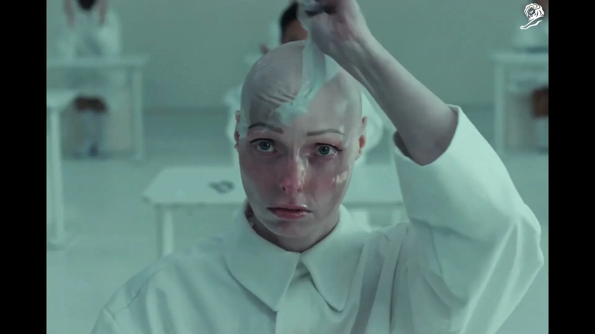 A bald woman in a white lab coat peels a clear mask from her face, looking directly at the camera in a sterile, futuristic lab.