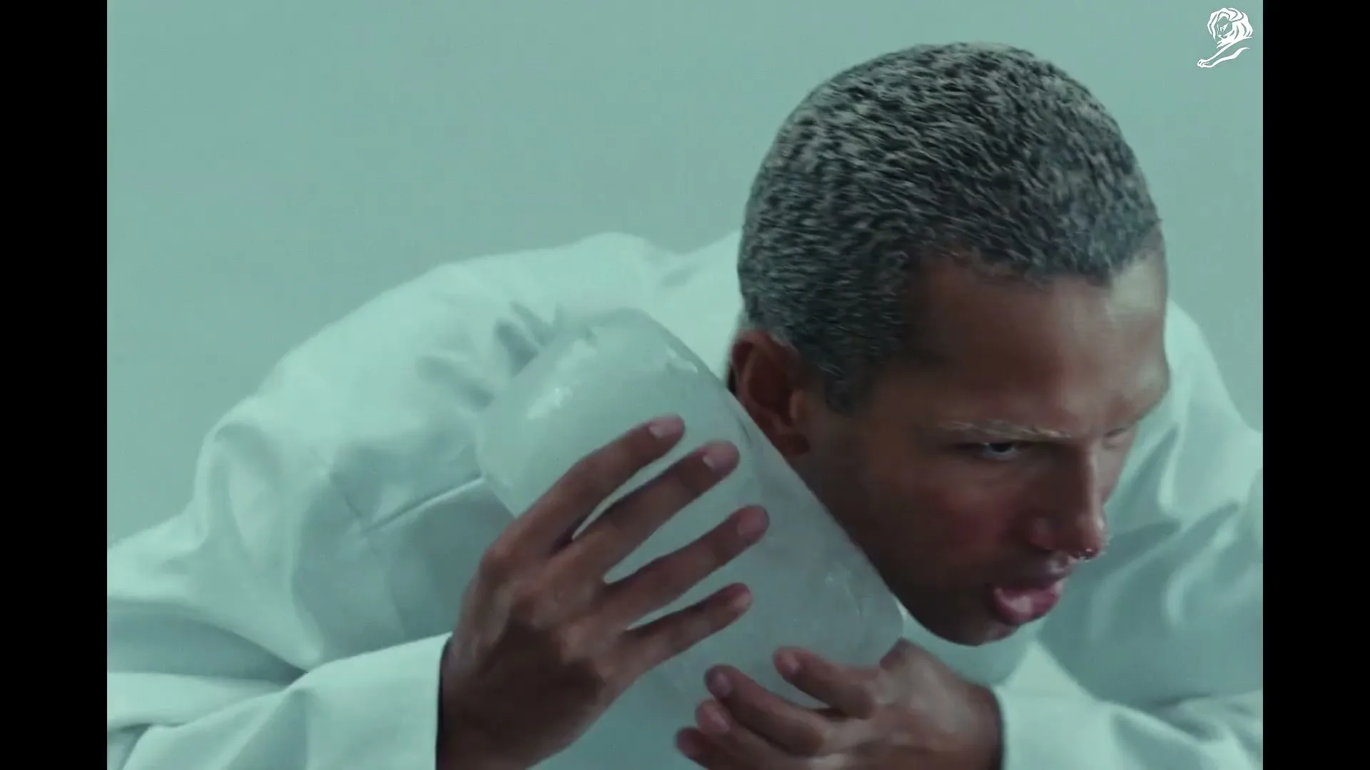A man with grey hair and a white lab coat holds a large piece of ice to his chest, looking distressed in a sterile, white room.