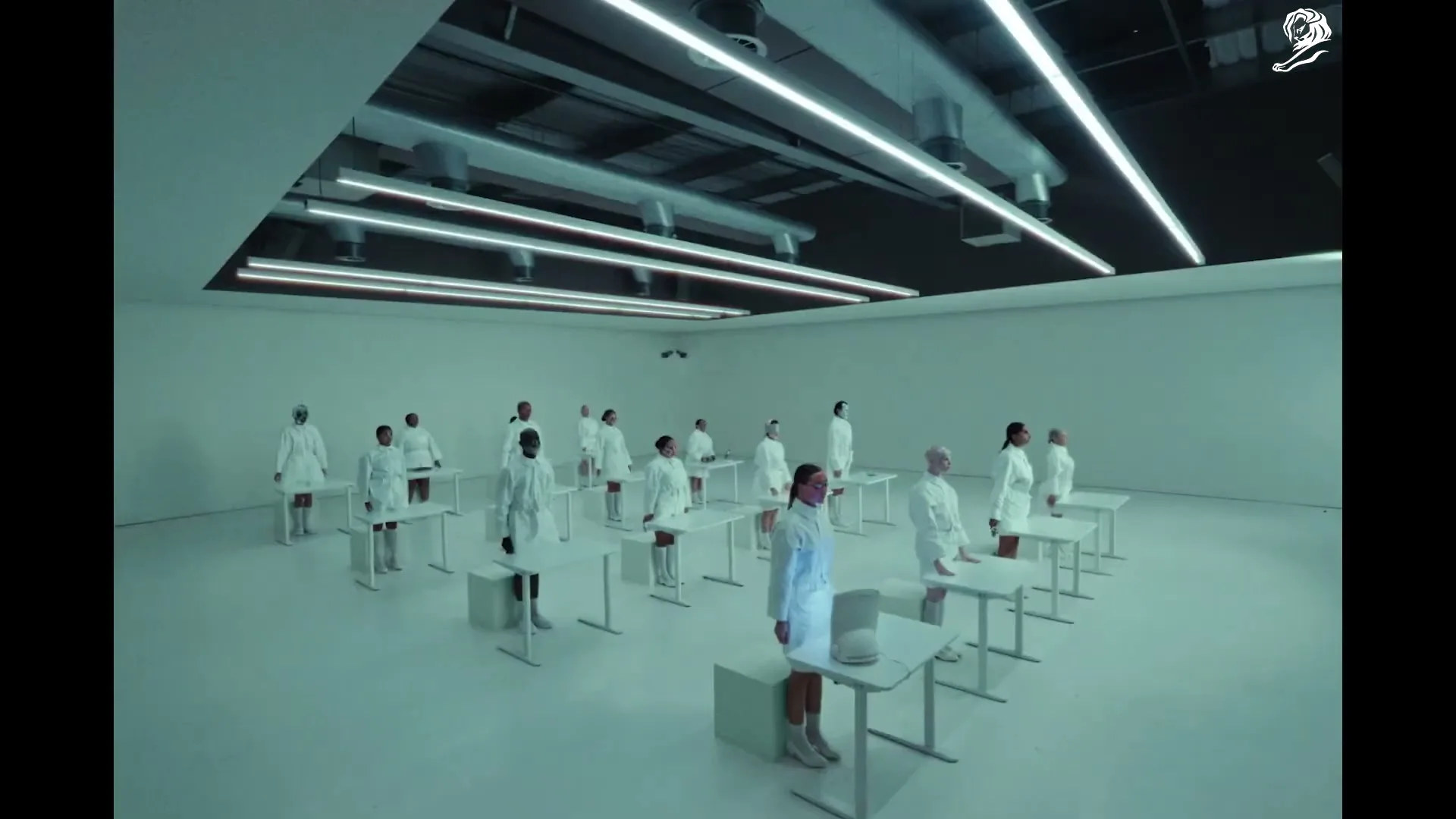 A group of diverse people in white lab coats stand behind individual white desks in a sterile, all-white room with bright overhead lights.