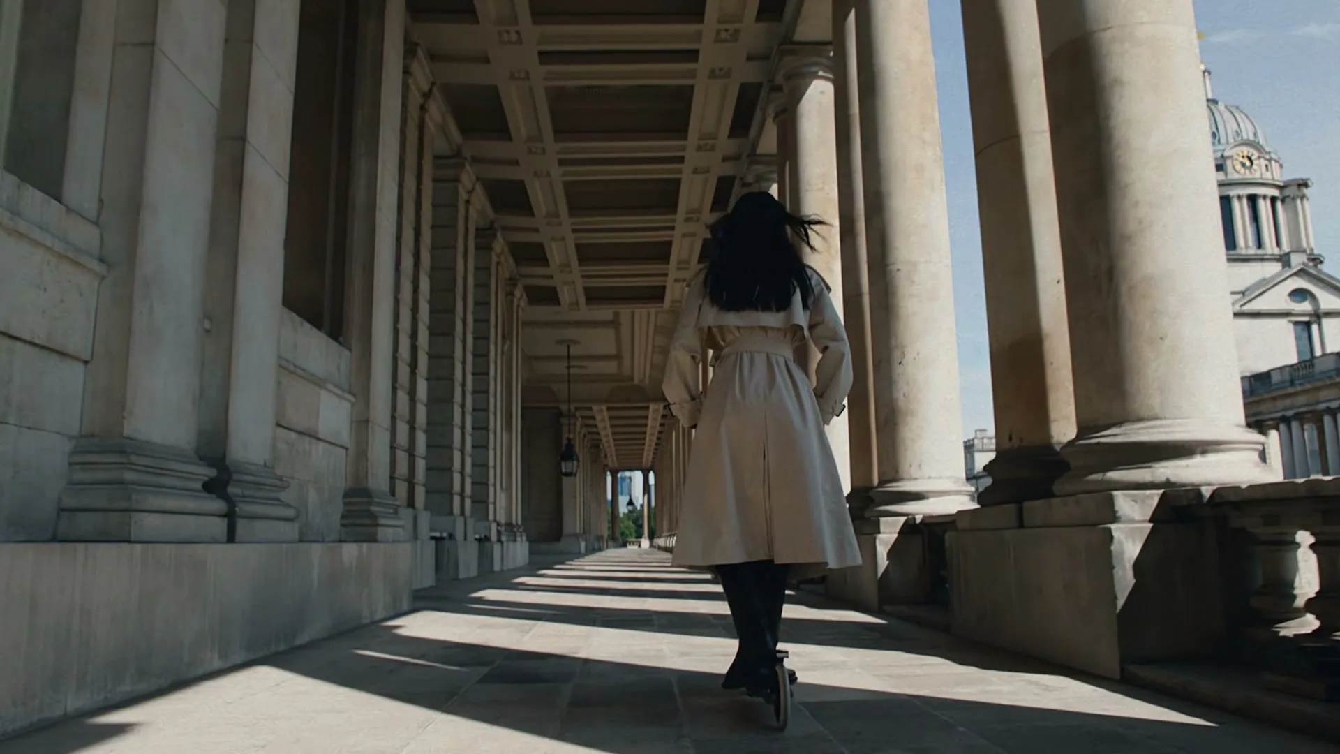 A woman with long dark hair, wearing a beige trench coat and black boots, rides a scooter down a long, columned hallway with a domed building in the background.