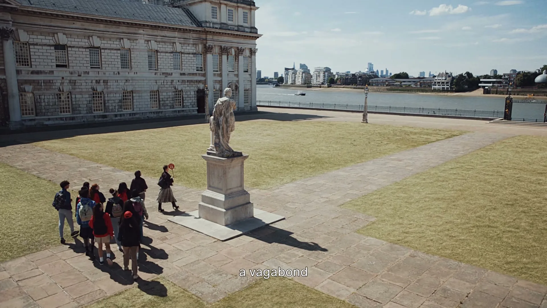 A group of young people walk past a statue in a courtyard next to a grand building, with a river and city skyline in the background on a sunny day.