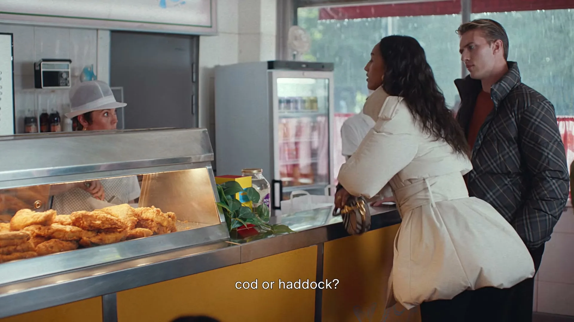 A young couple stands at a counter in a fish and chips shop, looking at the food and talking to the server.