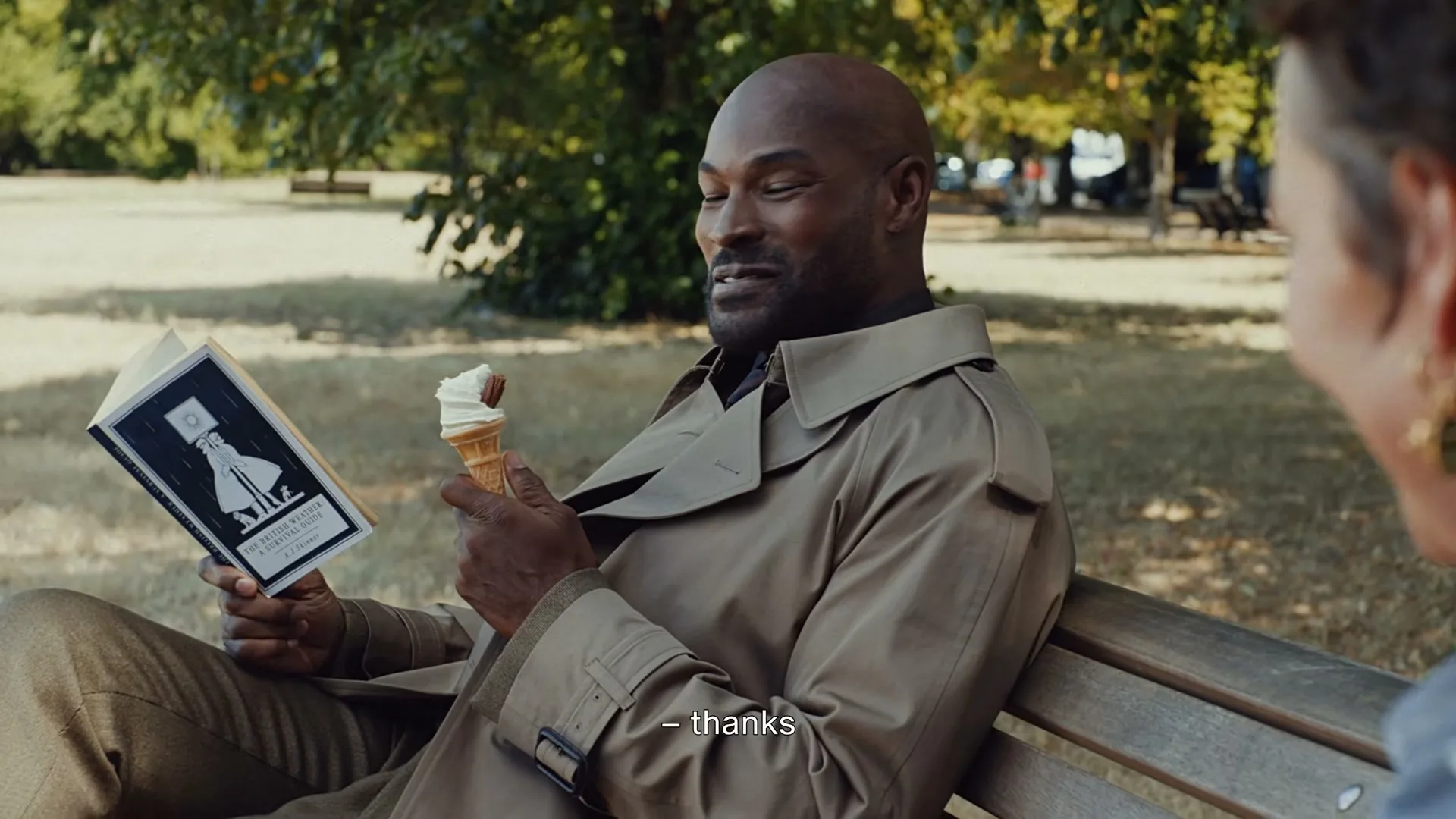A man in a trench coat sits on a park bench, reading a book and eating an ice cream cone, while talking to a woman.