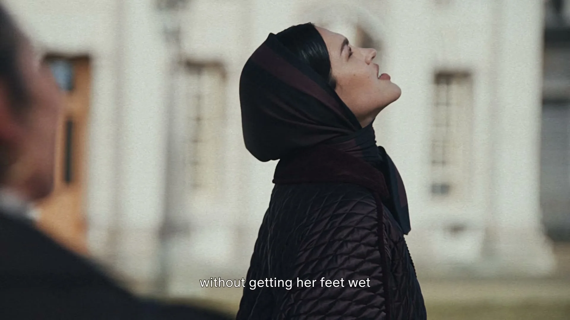 A woman in a dark quilted jacket and headscarf looks up at the sky, then turns to look at someone off-screen, outdoors in front of a building.