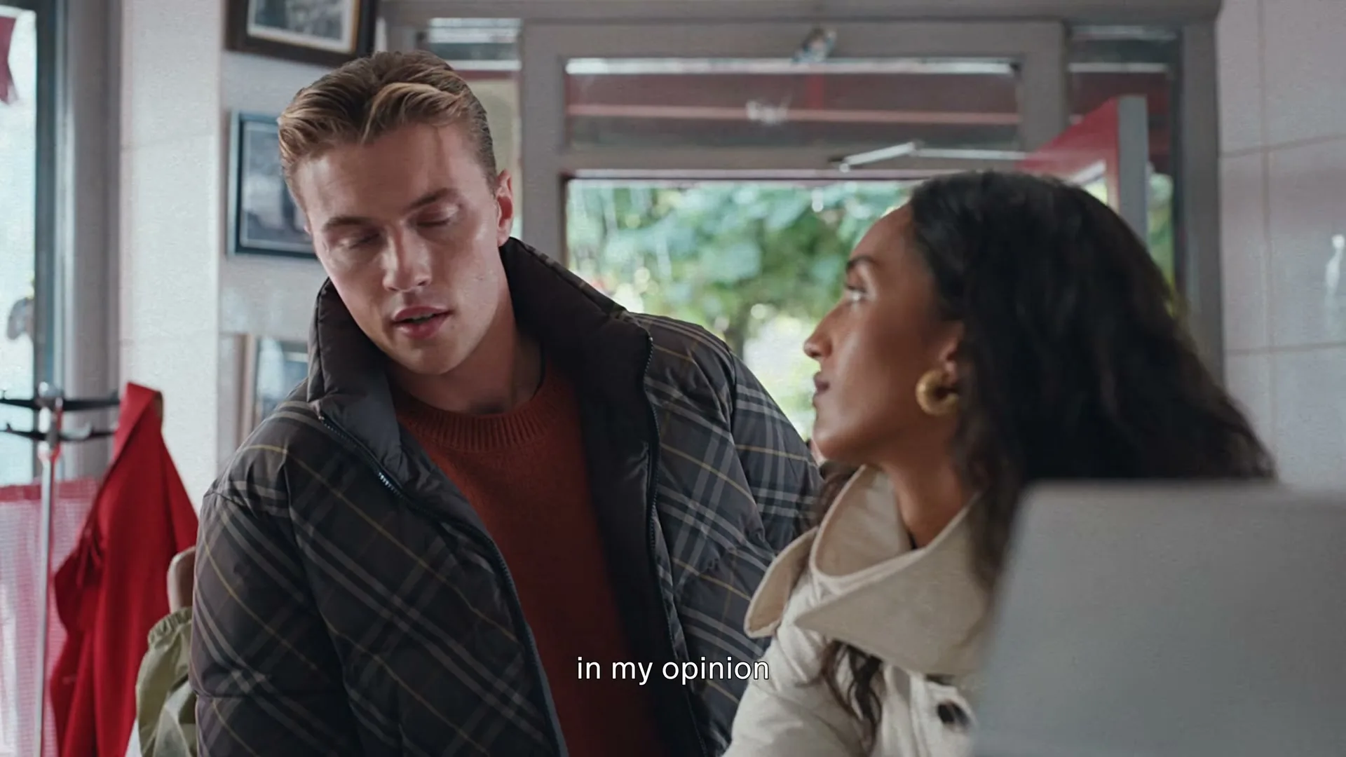 A young man in a plaid puffer jacket looks at a young woman in a cream coat, both standing inside a fish and chips shop.