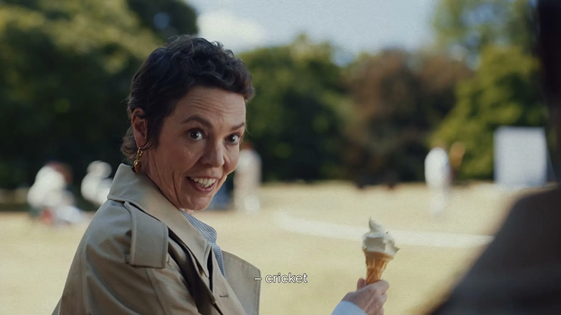 A woman in a trench coat and striped shirt smiles at the camera while holding an ice cream cone in a park where people are playing cricket.