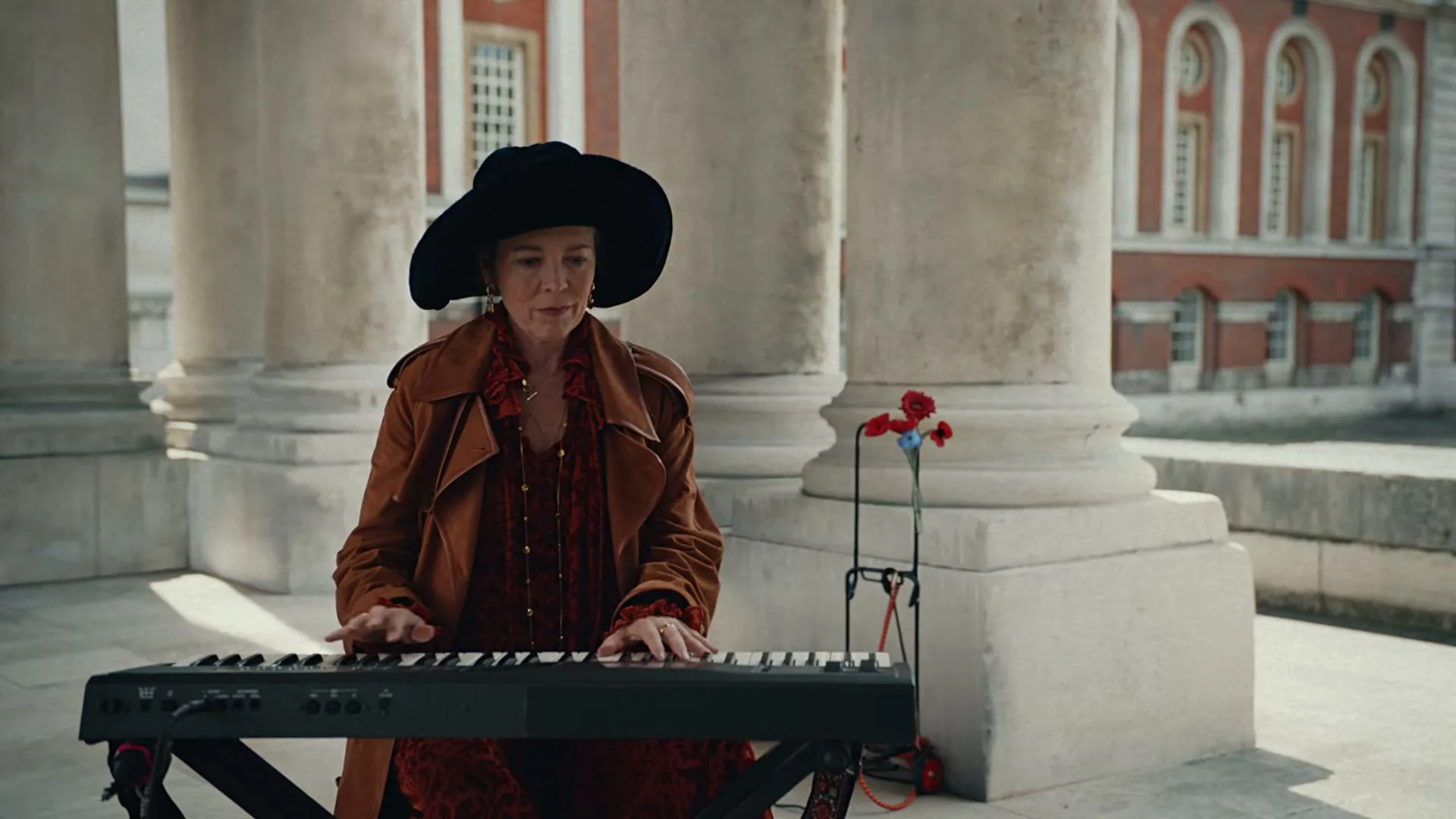 A woman in a wide-brimmed hat and trench coat plays a keyboard under a colonnade, with a red brick building in the background.