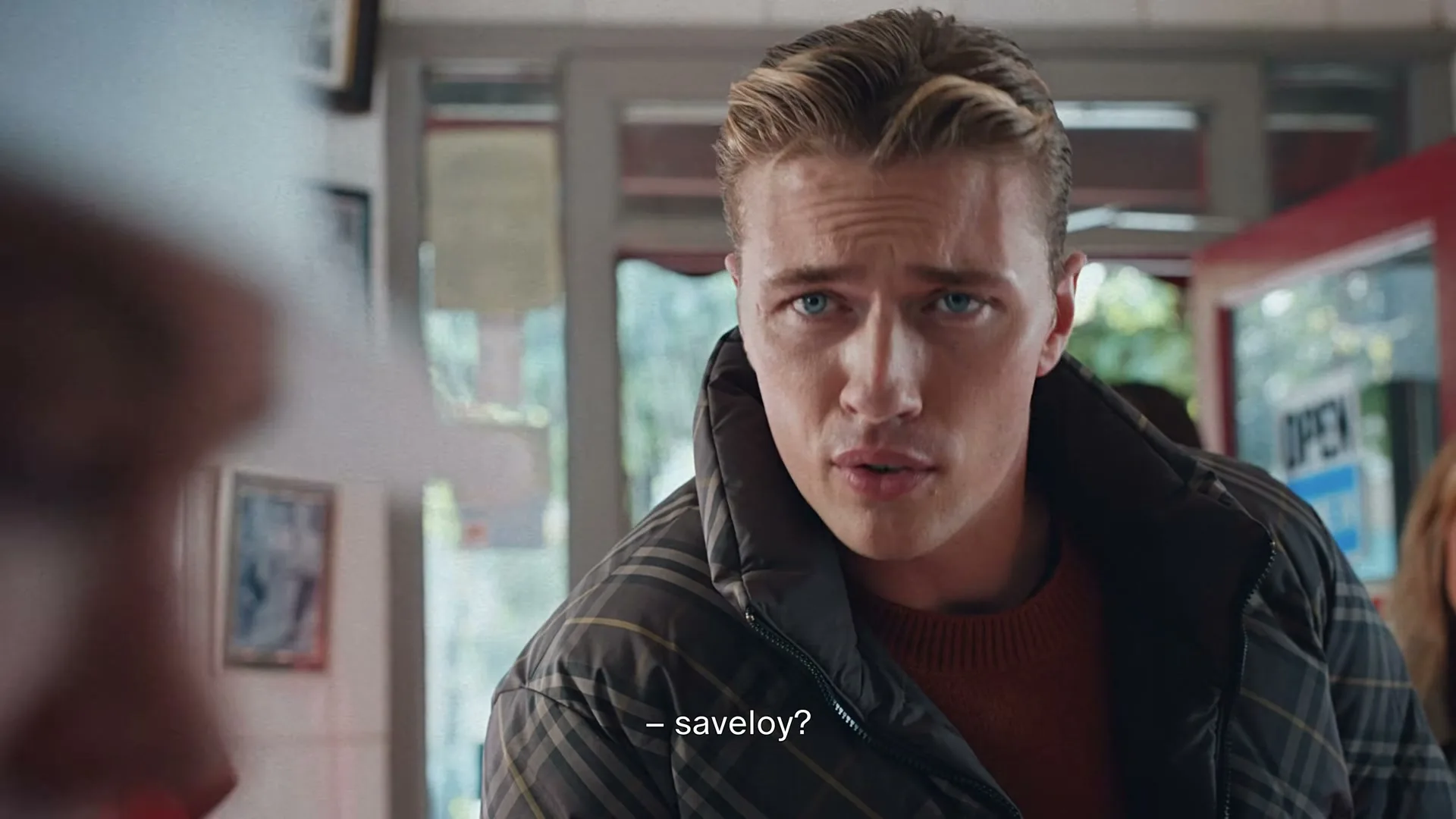 A young man with blonde hair and blue eyes, wearing a plaid jacket, looks intently at someone off-screen in a shop.