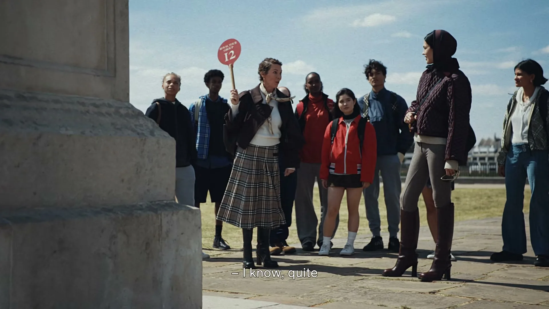 A diverse group of people stands outdoors on a sunny day, watching a woman in a plaid skirt hold up a red sign with the number 12.