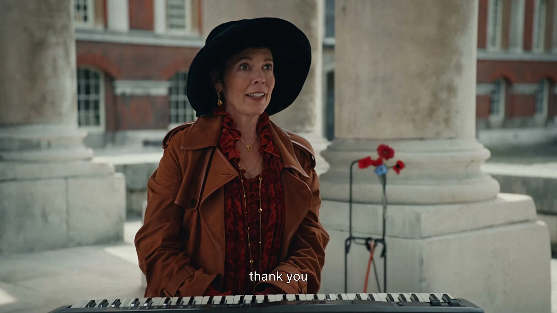 A woman in a black hat and brown trench coat plays a keyboard outdoors, looking up and smiling at someone off-camera.