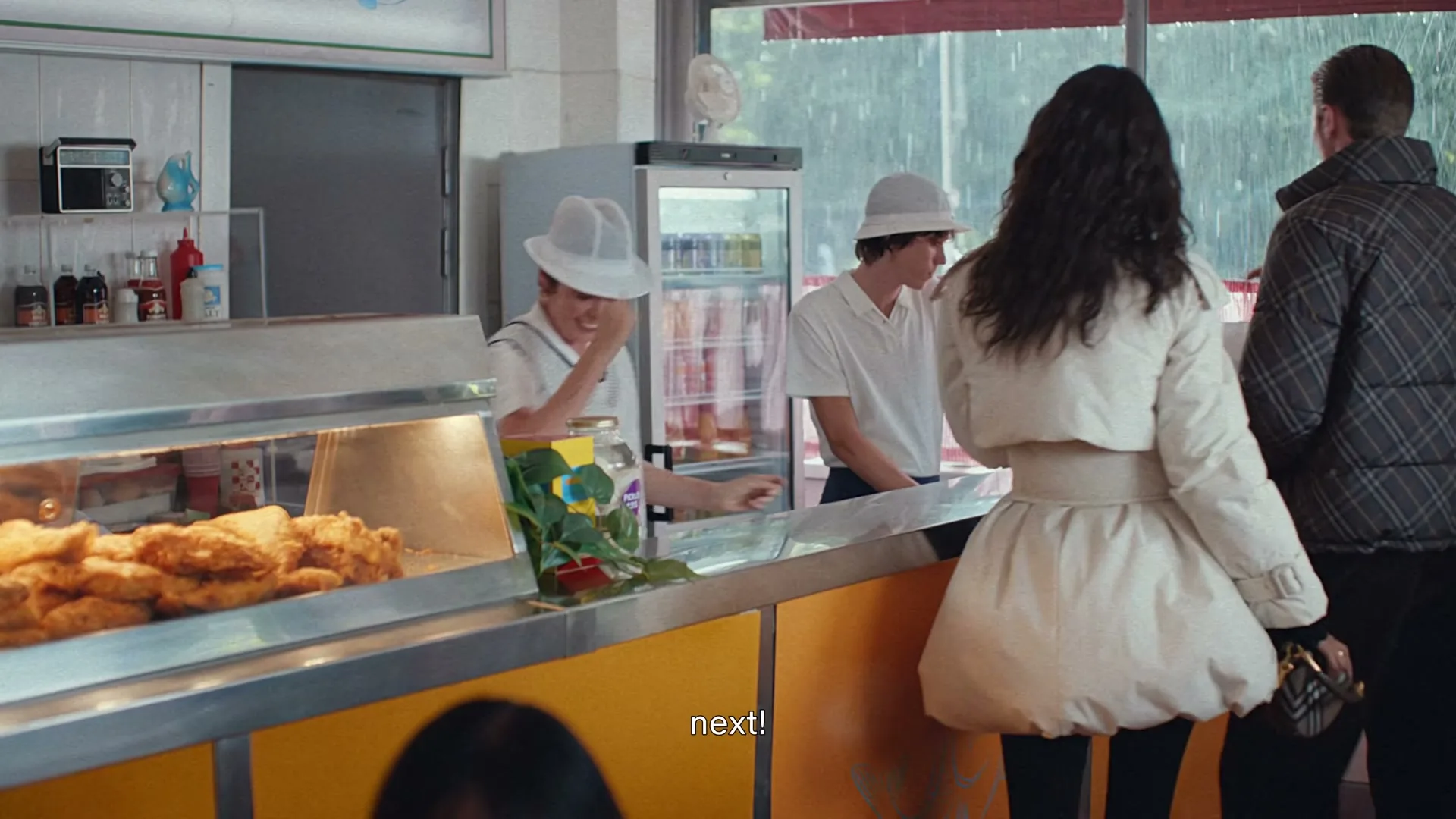 A woman in a white puffer jacket stands at a counter in a fish and chips shop, while a server smiles and gestures to the next customer.