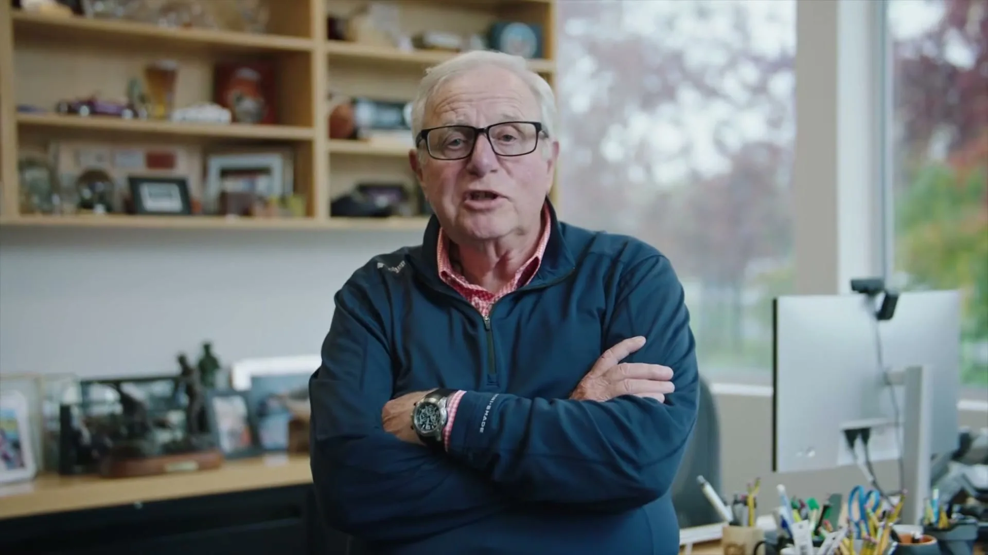 Tim Boyle, CEO of Columbia Sportswear, a 60s+ male with white hair and glasses, stands with crossed arms in his office, speaking directly to the camera.