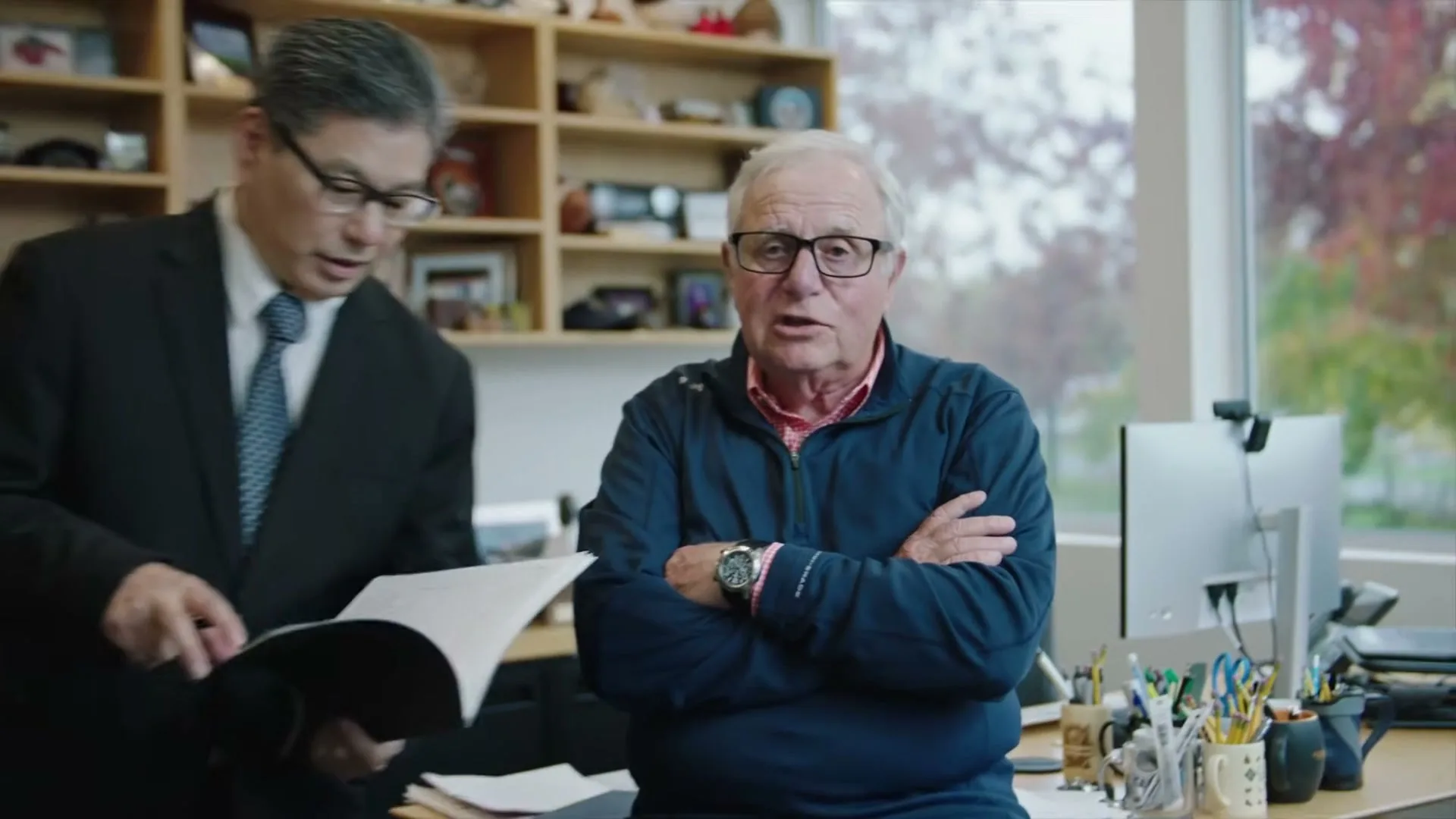 An older man in glasses and a blue jacket speaks to the camera, then an Asian man in a suit walks in holding paperwork in an office.