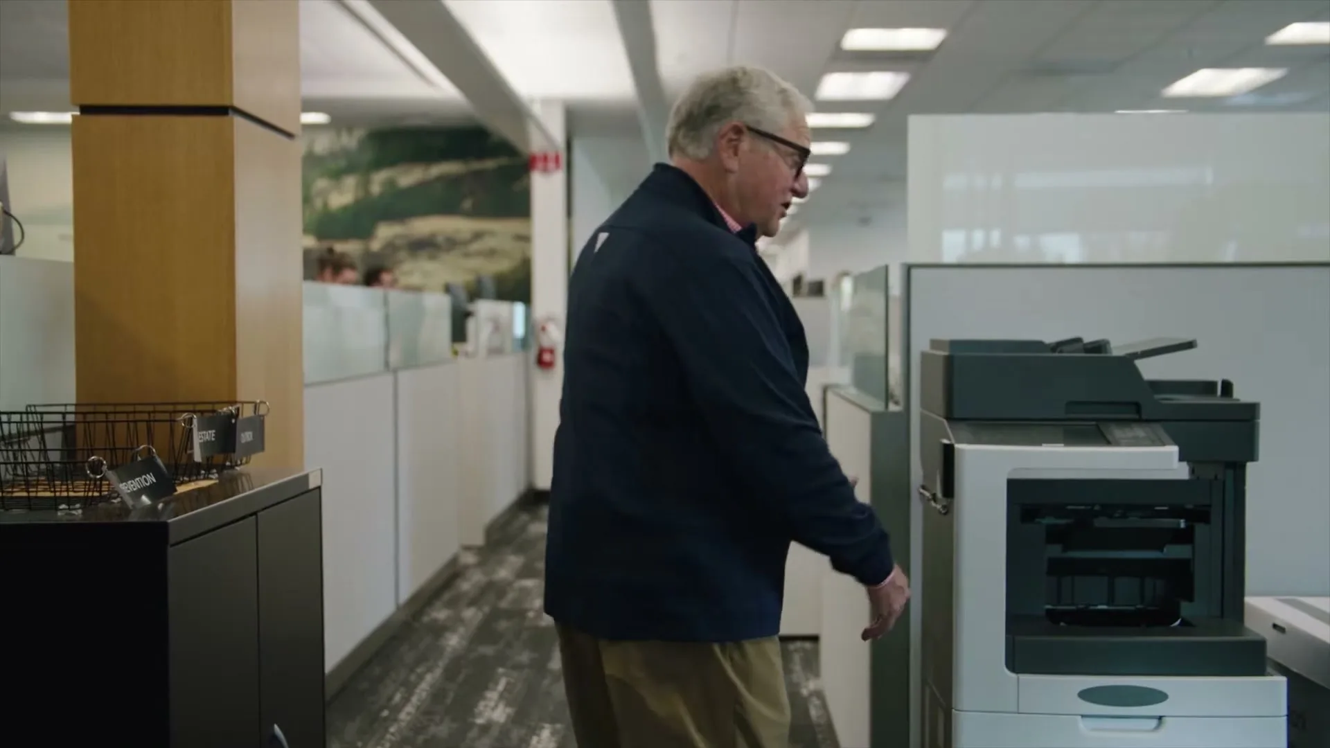 An older man in glasses and a navy jacket walks past a printer in a modern office hallway, looking back over his shoulder.