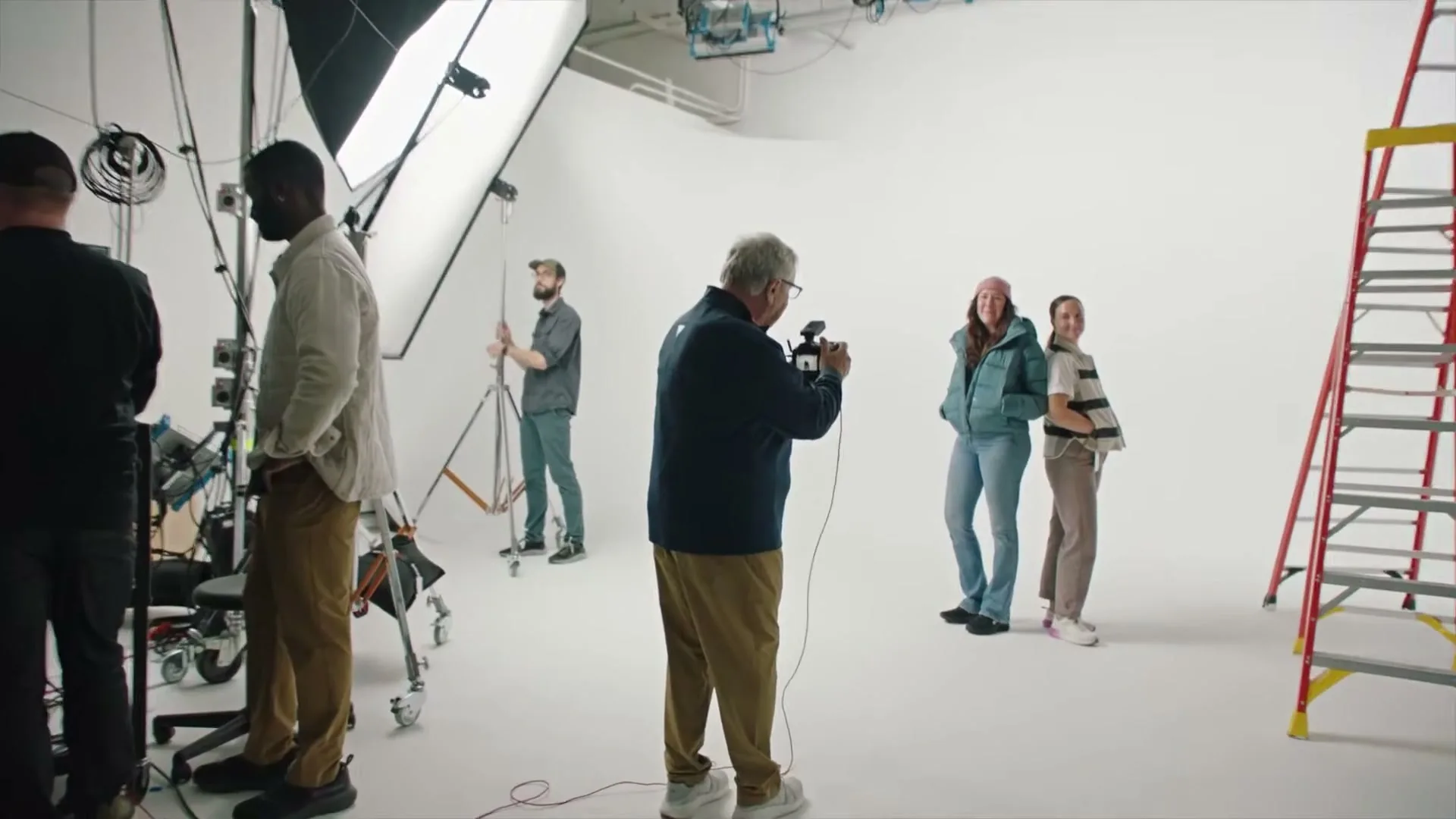 A male photographer in a blue jacket takes photos of two female models in winter clothing in a brightly lit studio with crew and equipment.