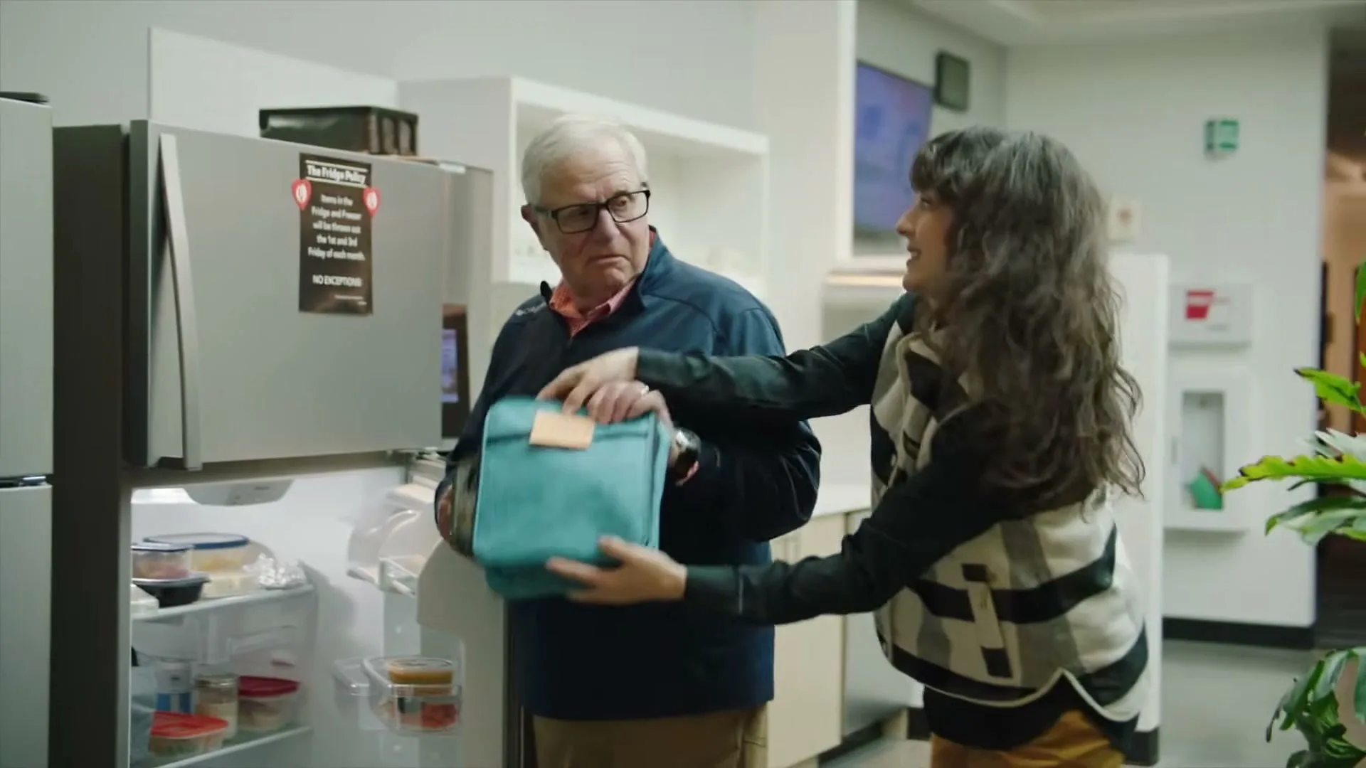 An older man in glasses and a younger woman in a striped vest interact by an open refrigerator in a modern office kitchen.