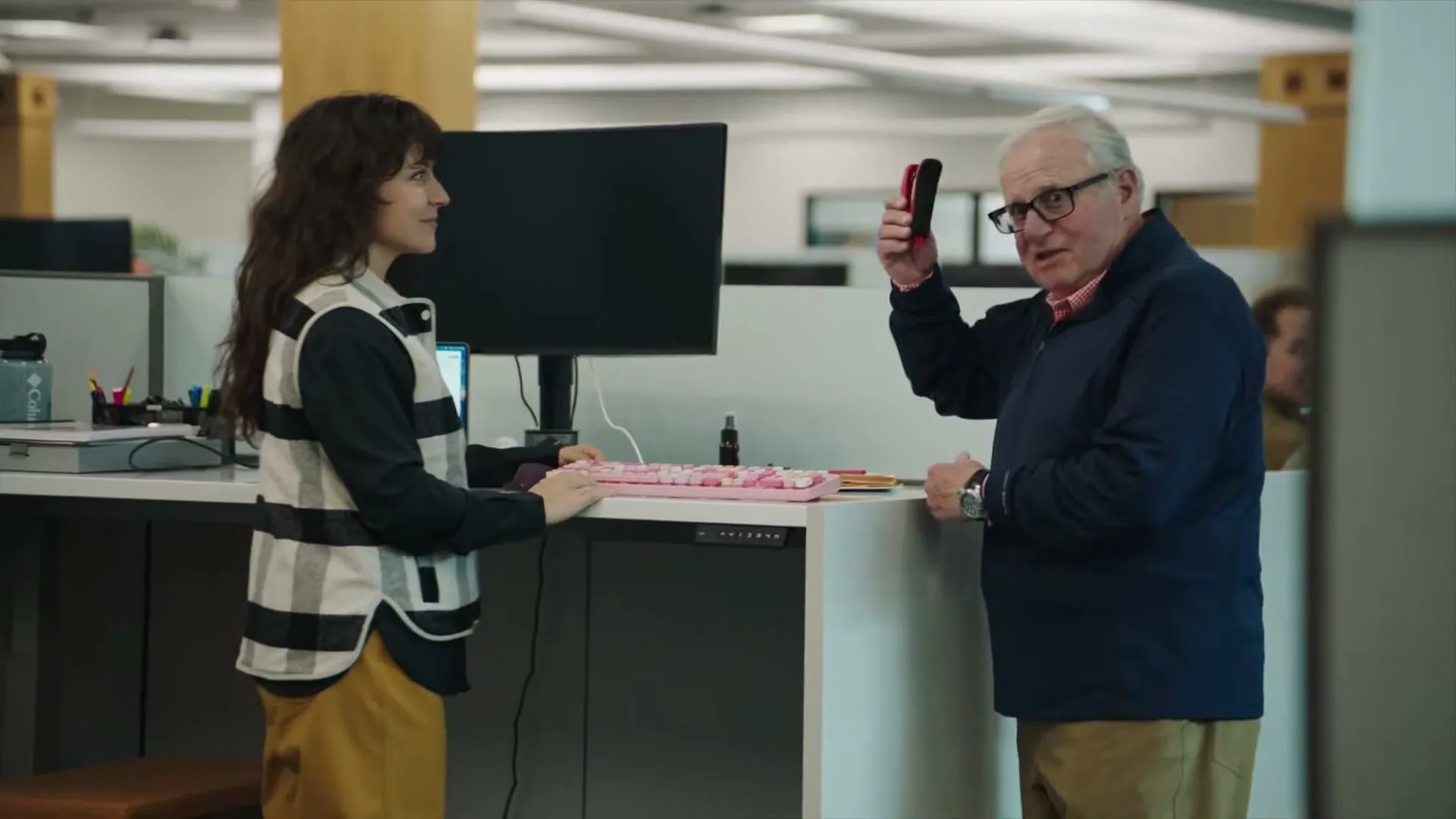 A young woman with dark curly hair and an older man with white hair stand at an office desk, the man holding up a small red and black device.