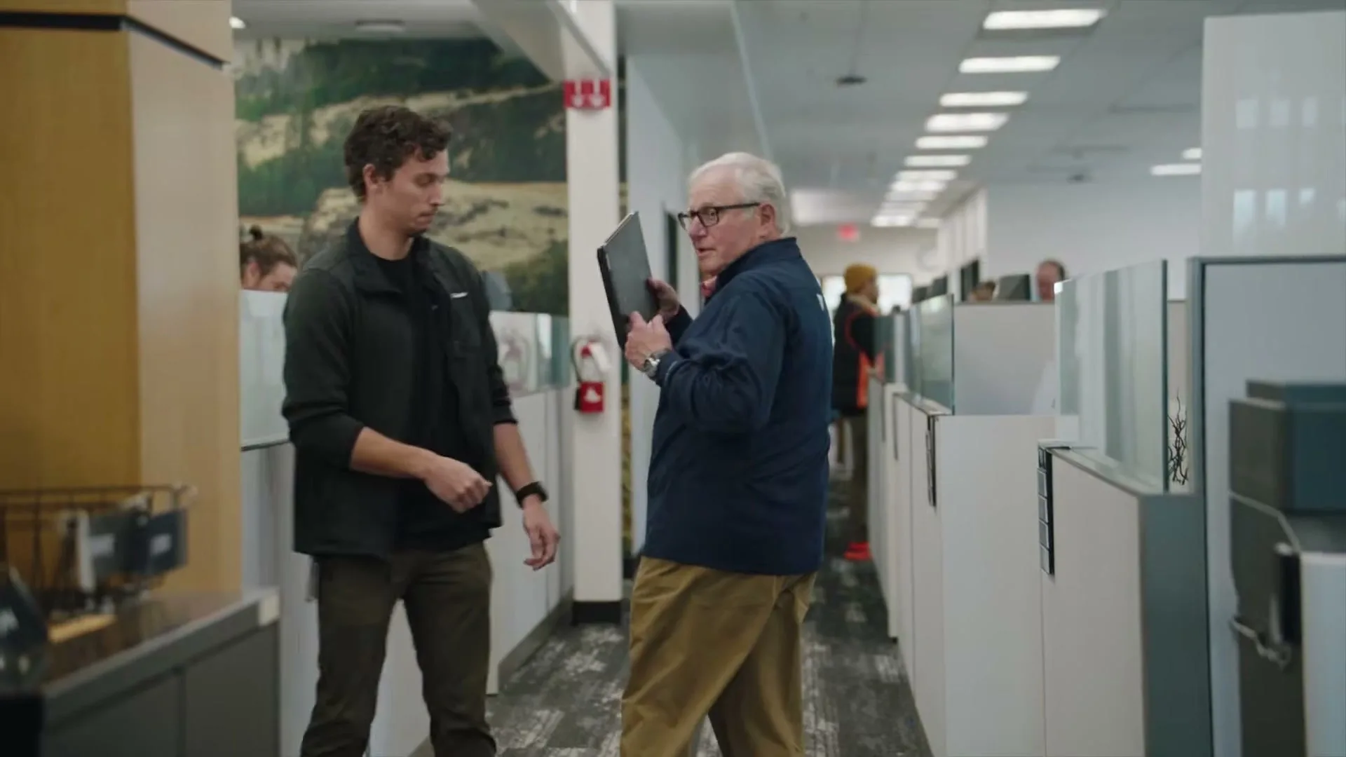 A young man in a dark jacket and an older man in a blue shirt and khakis walk through an office hallway, exchanging a laptop.