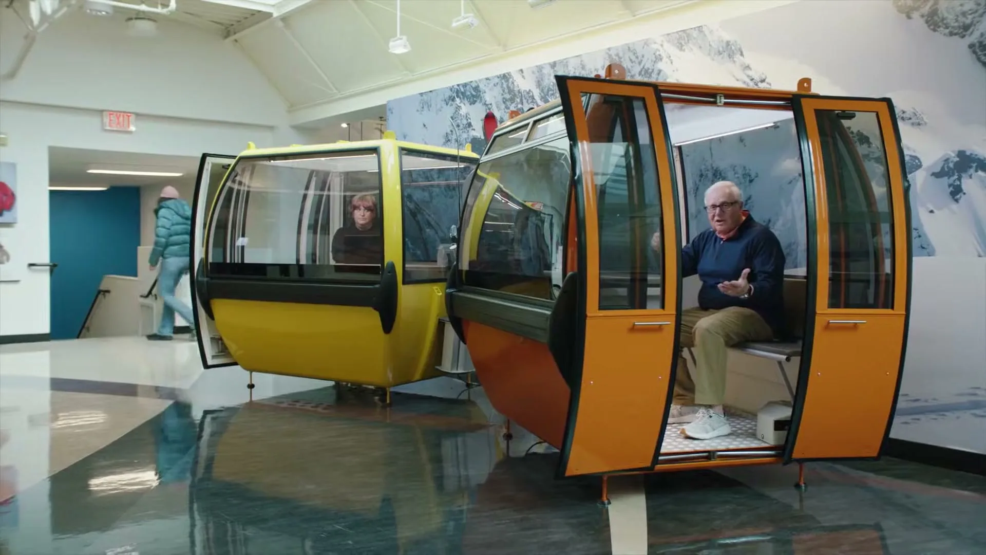 An older man and a younger woman sit in separate gondola cars inside a building with a mountain mural, while another person walks by.