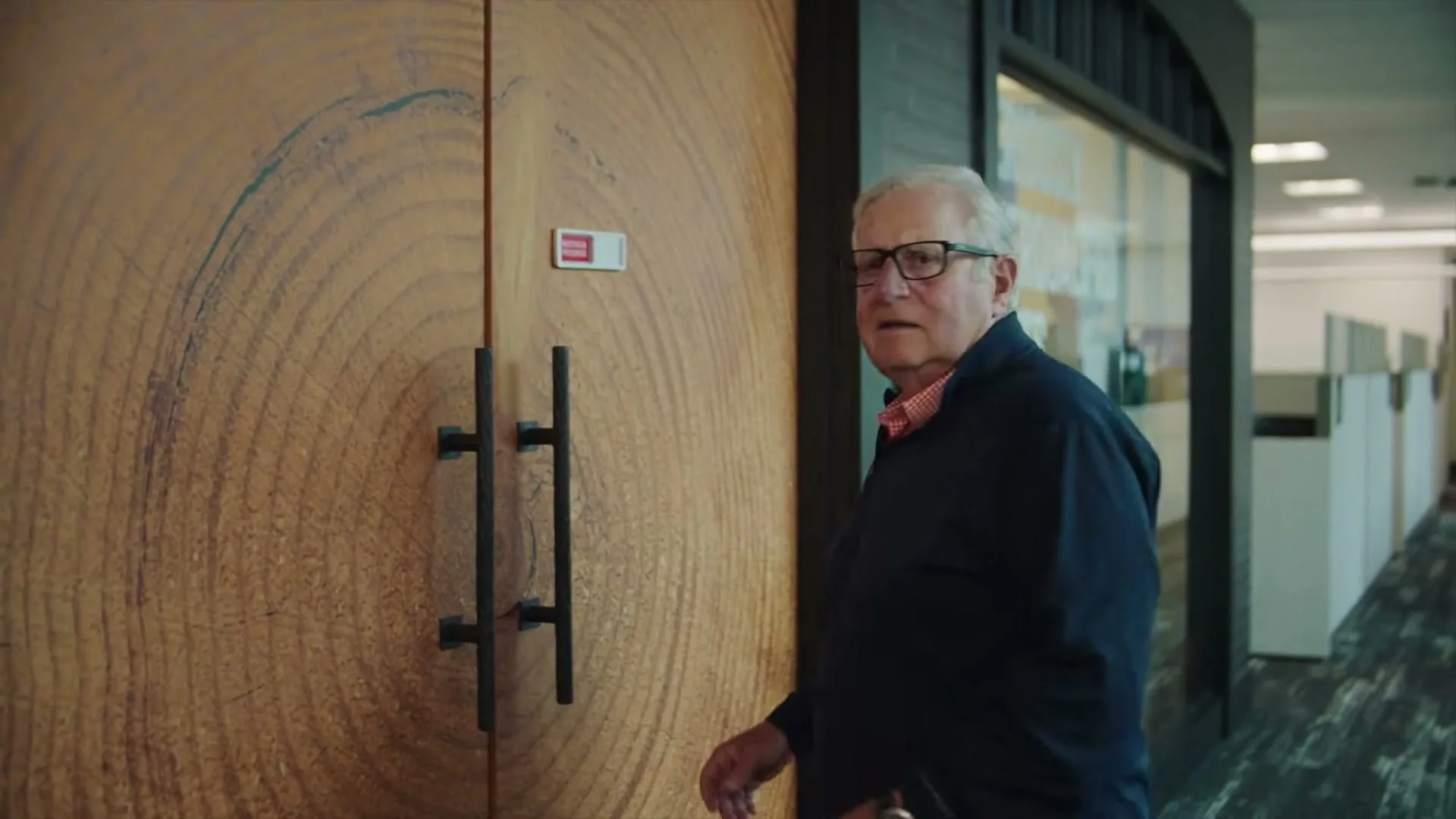 An older white man with glasses and a blue jacket walks away from a wooden door, then turns to look back with a confused expression in an office hallway.