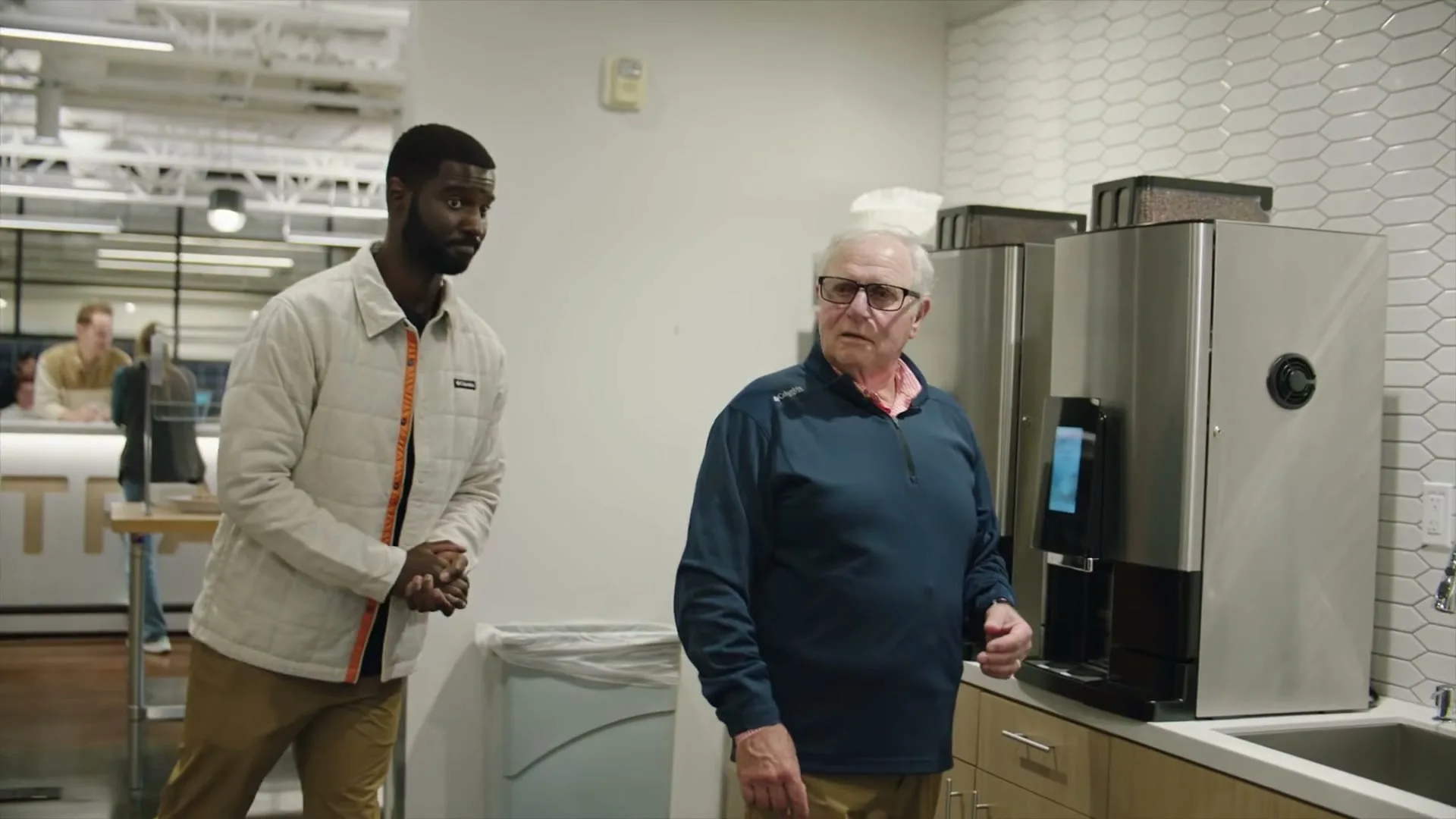 An older man in glasses and a blue jacket stands at a coffee machine in an office kitchen, while a younger man in a white jacket approaches him.