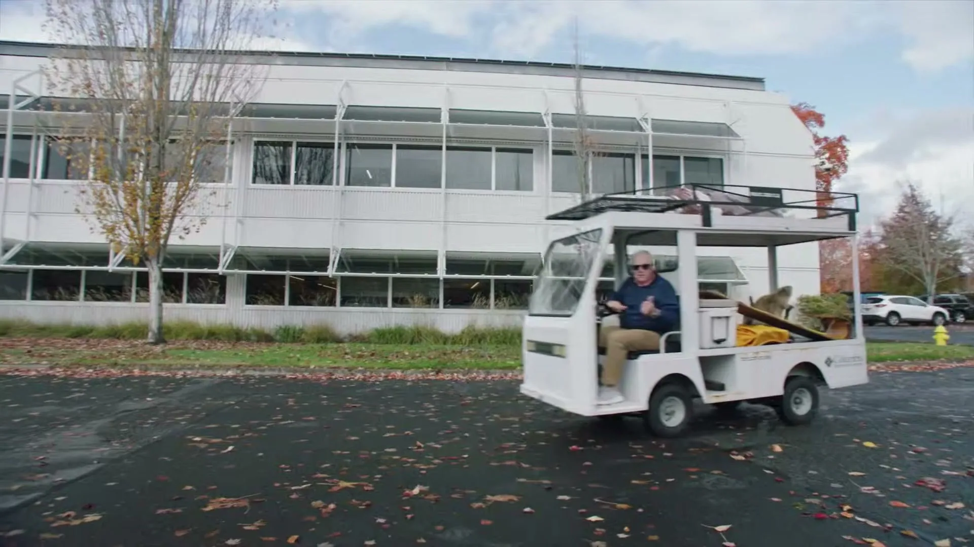 A man in his 60s drives a small white utility vehicle with camping gear past a modern office building on a cloudy day.